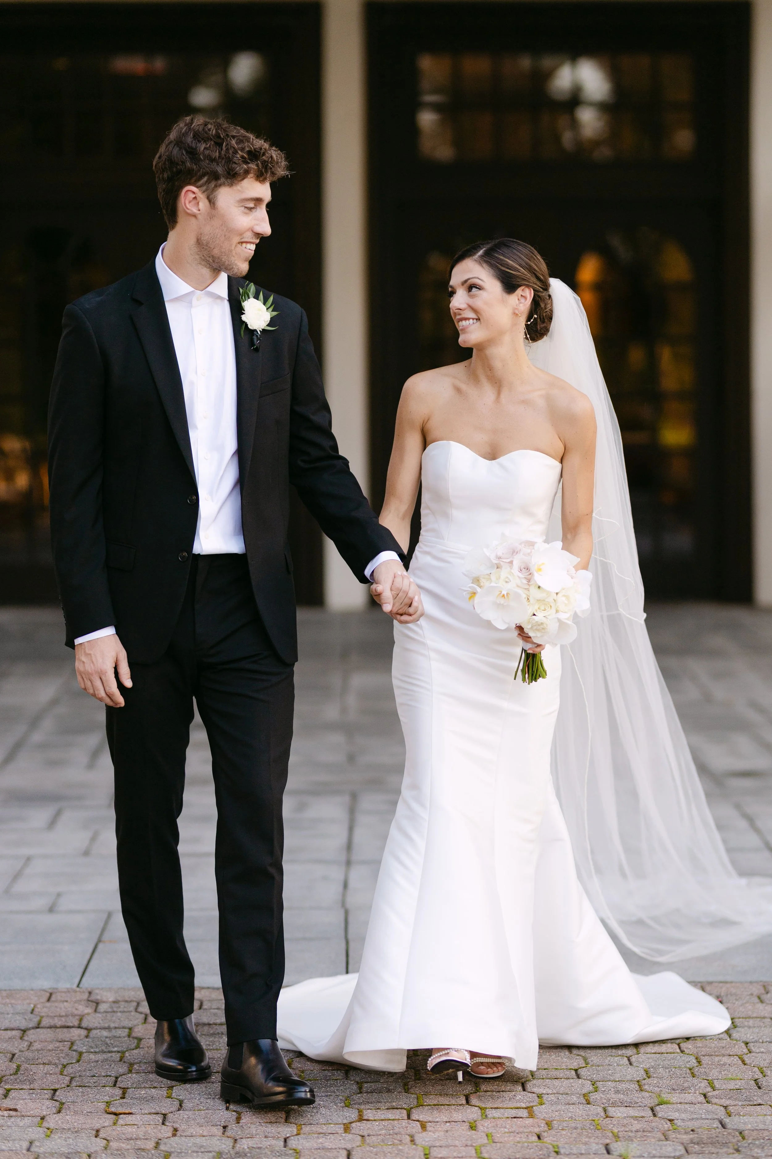 A bride and groom holding hands and smiling at each other outside a building, dressed in wedding attire, with the bride holding a bouquet of white flowers.