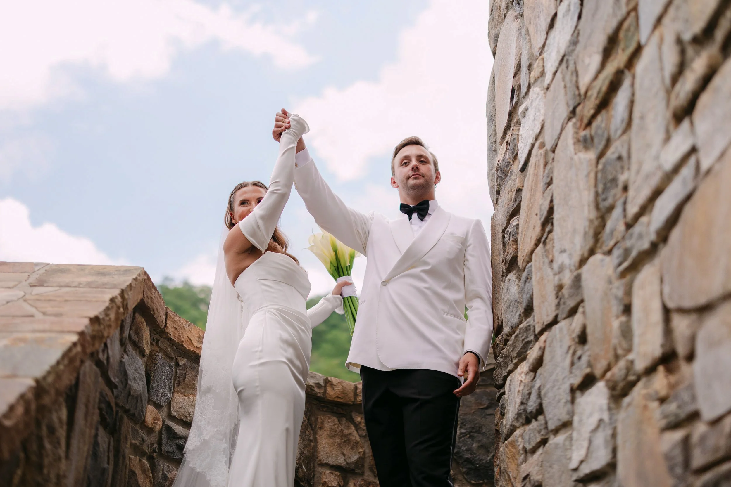 A bride and groom holding hands and walking together outdoors. The bride is holding a bouquet of white calla lilies. The scene is set against a stone wall with a cloudy sky.