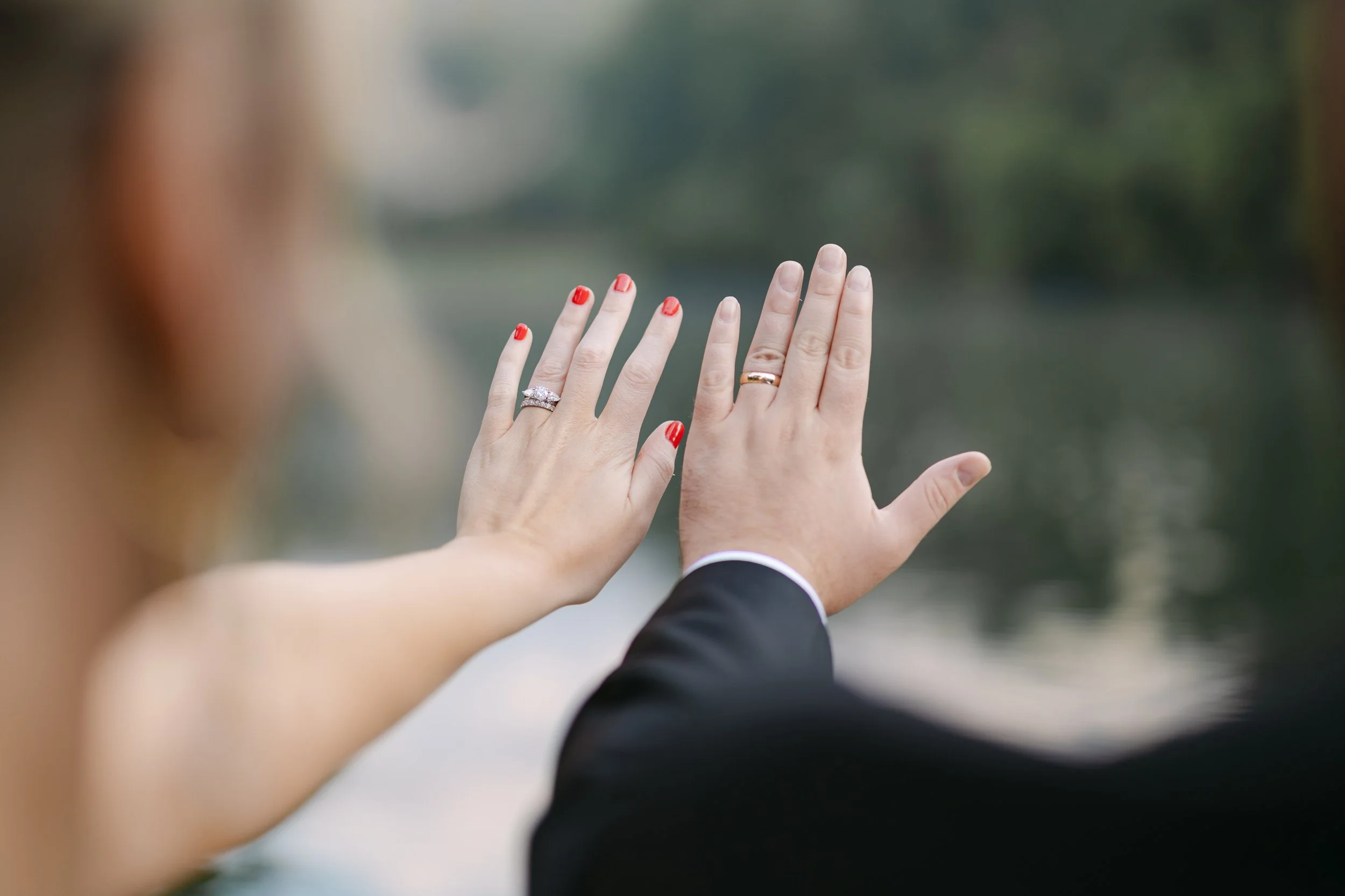 A bride and groom showing their wedding rings with one hand each, against a blurred outdoor background.