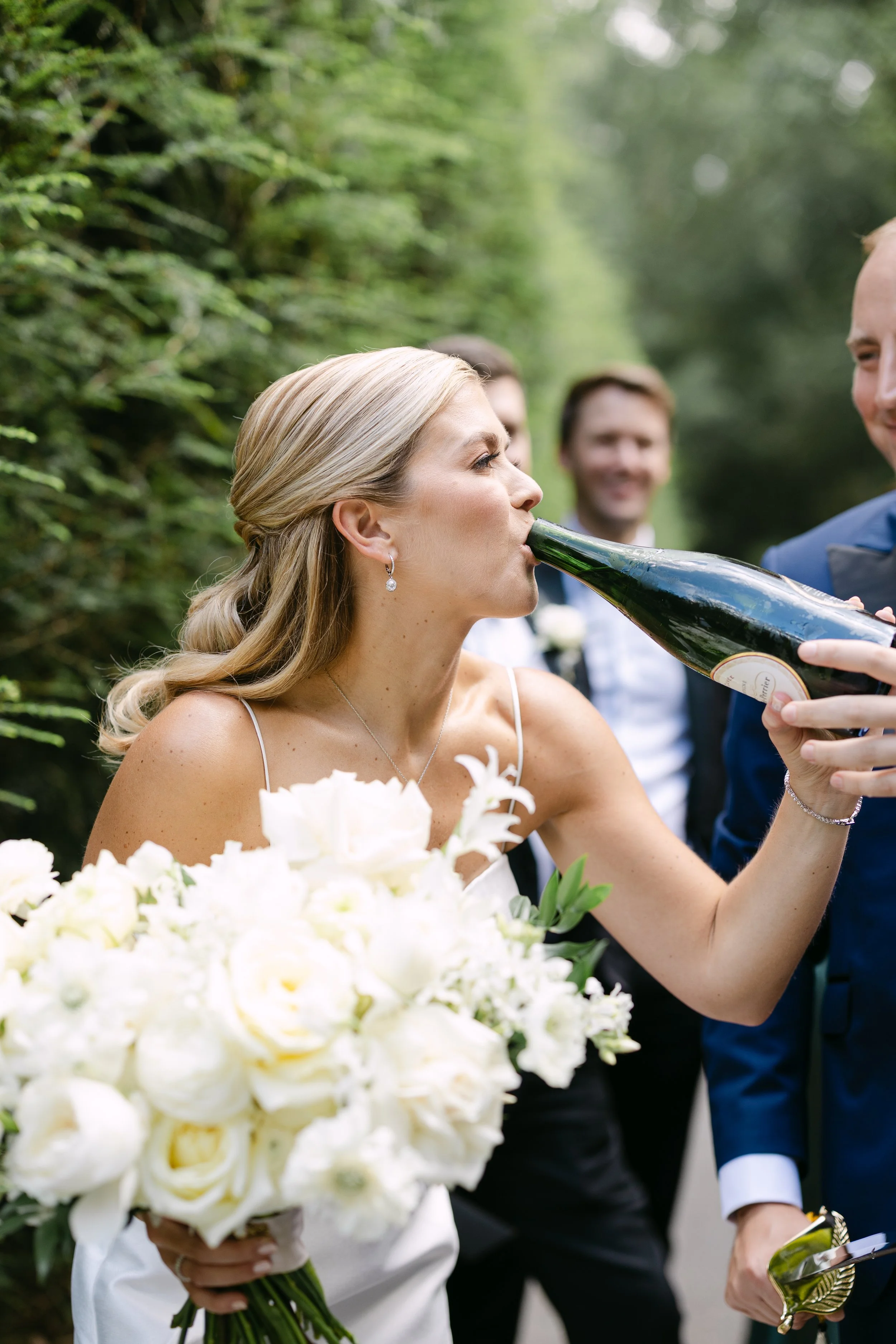 Bride in wedding dress holding a bouquet of white flowers drinks champagne from a bottle held by groom during outdoor wedding celebration.