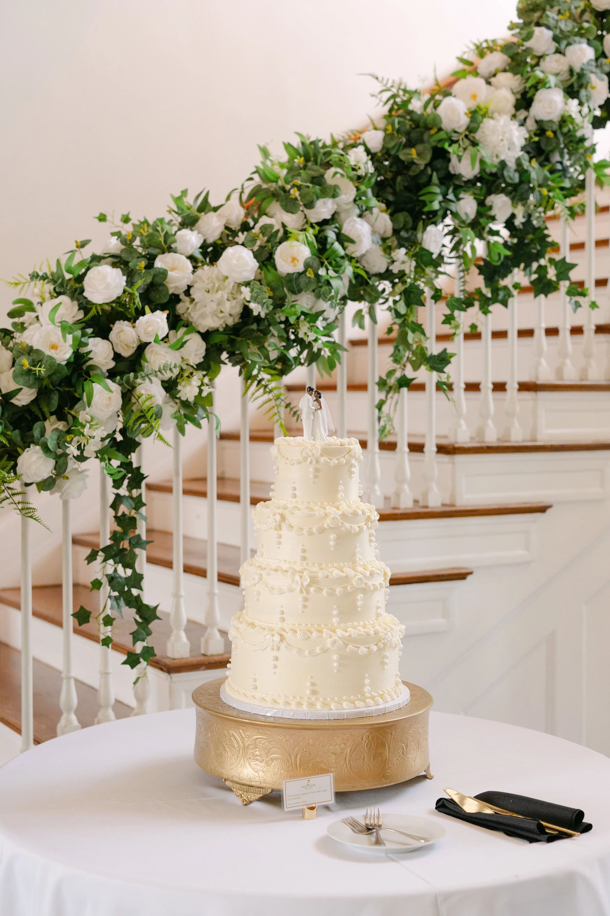 Wedding cake with four tiers decorated with white icing and pearl-like accents, topped with a bride and groom figurine. The cake is on a gold stand on a round table covered with a white tablecloth, next to a plate, fork, and napkin. In the background
