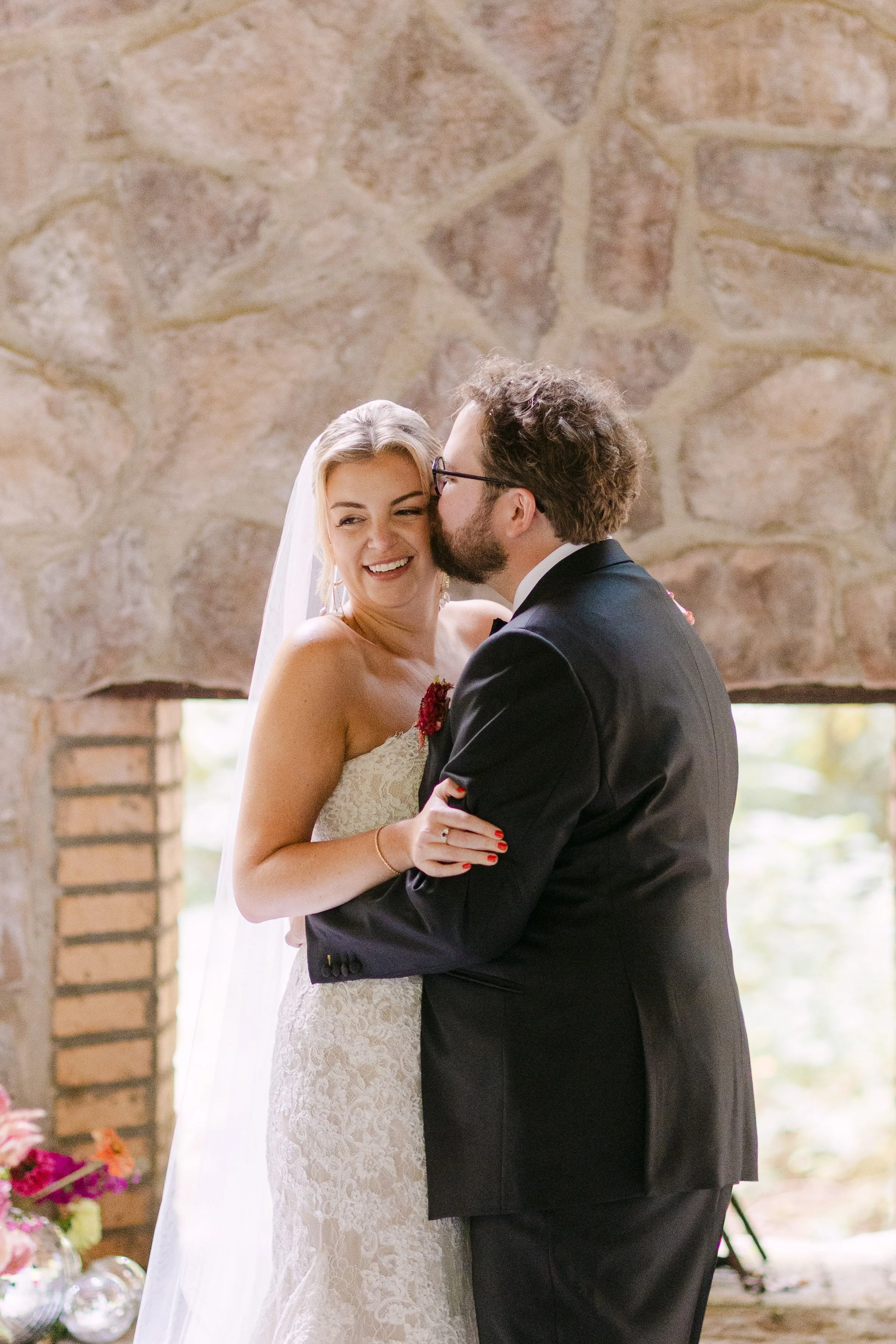 A bride and groom sharing a kiss indoors, with the bride smiling, near a stone wall background.
