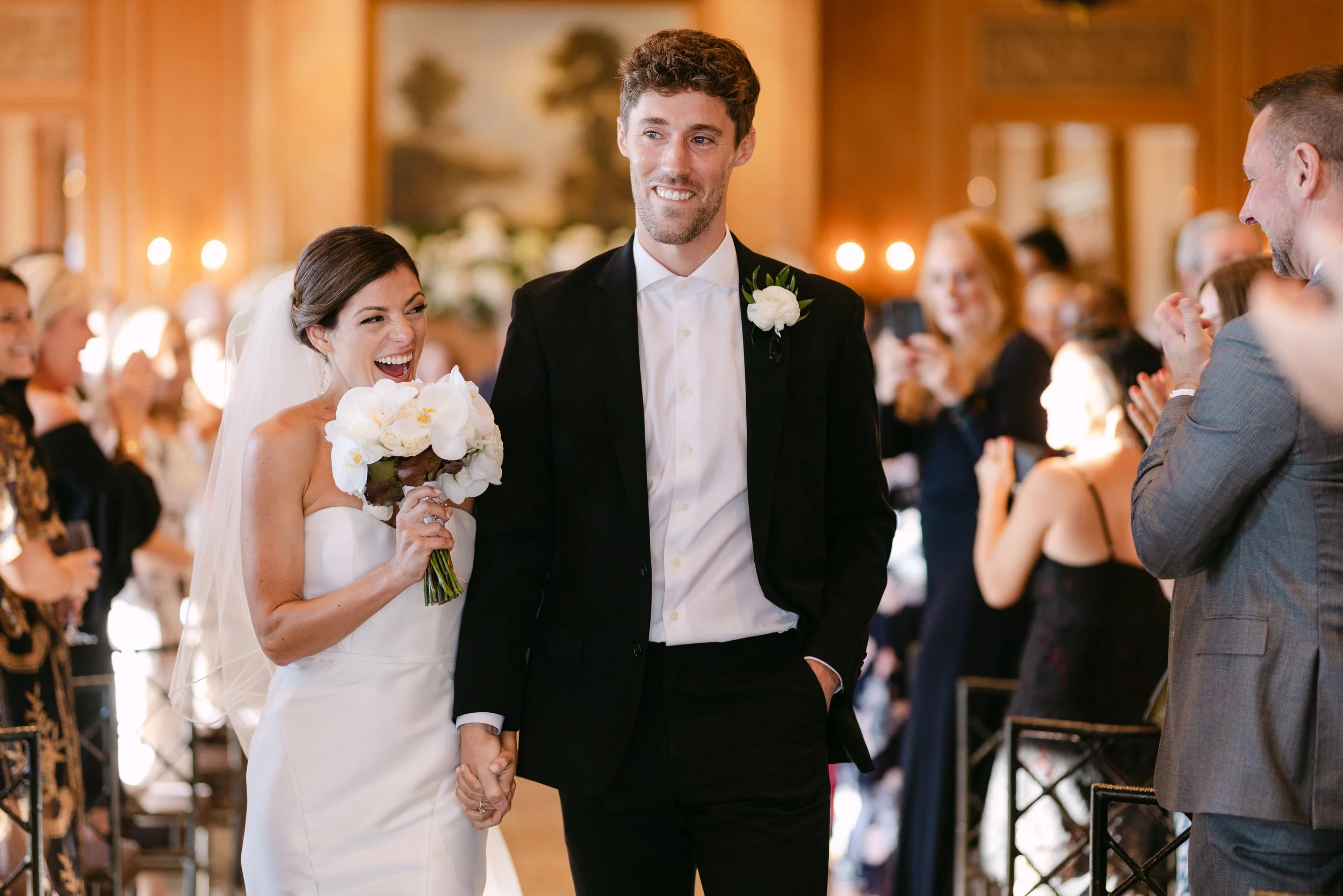 Bride and groom holding hands, smiling at each other during their wedding ceremony, with guests clapping and taking photos in the background.