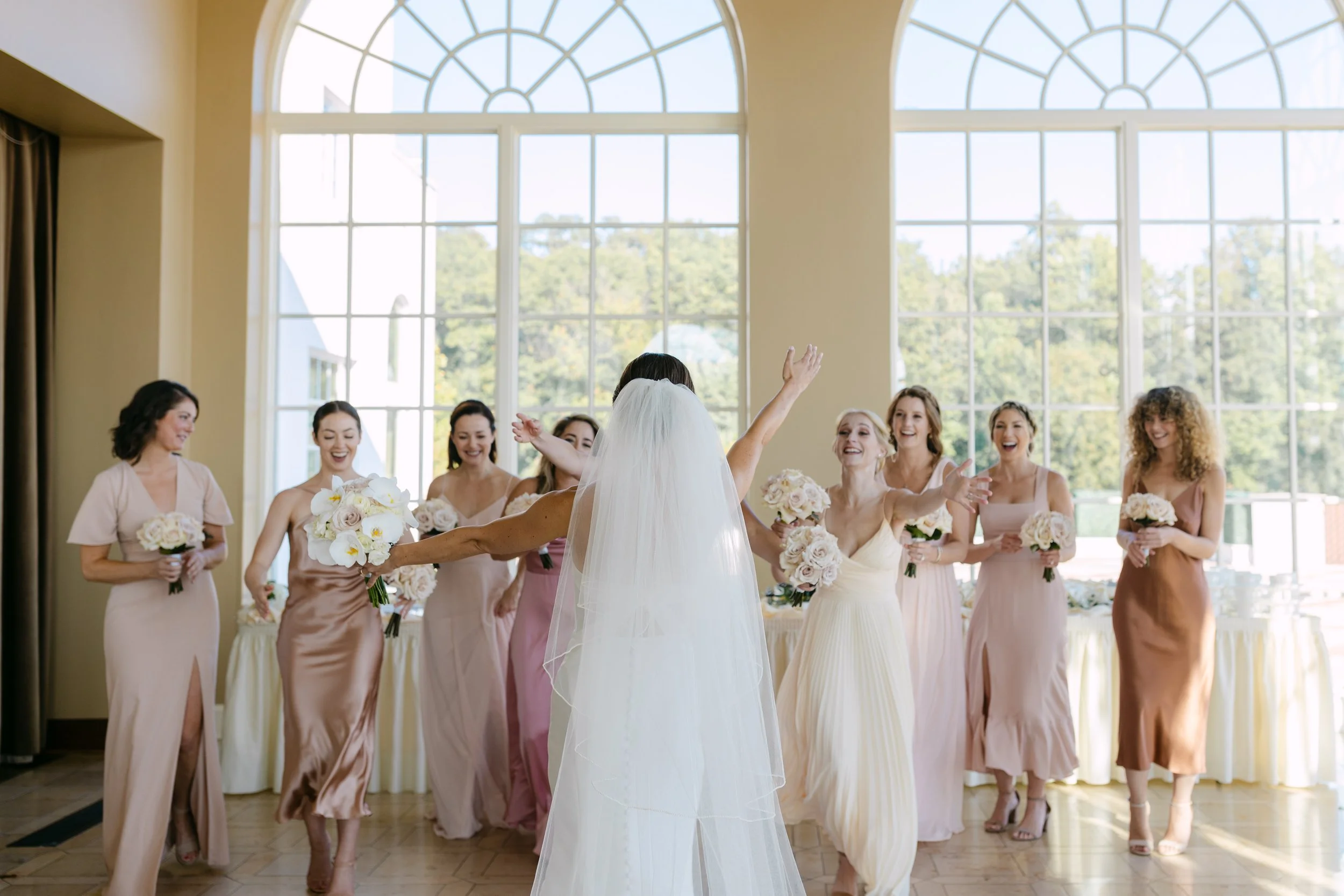 Bride in white wedding gown and veil with arms open, surrounded by bridesmaids holding bouquets, in a bright room with large windows.