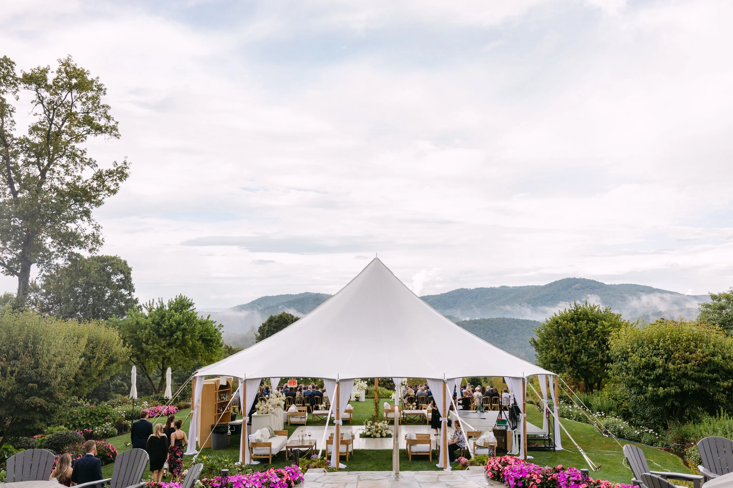 Wedding reception outdoors under a large white tent, with guests seated at tables on a lush green lawn, surrounded by flowers and trees, with mountains and cloudy sky in the background.