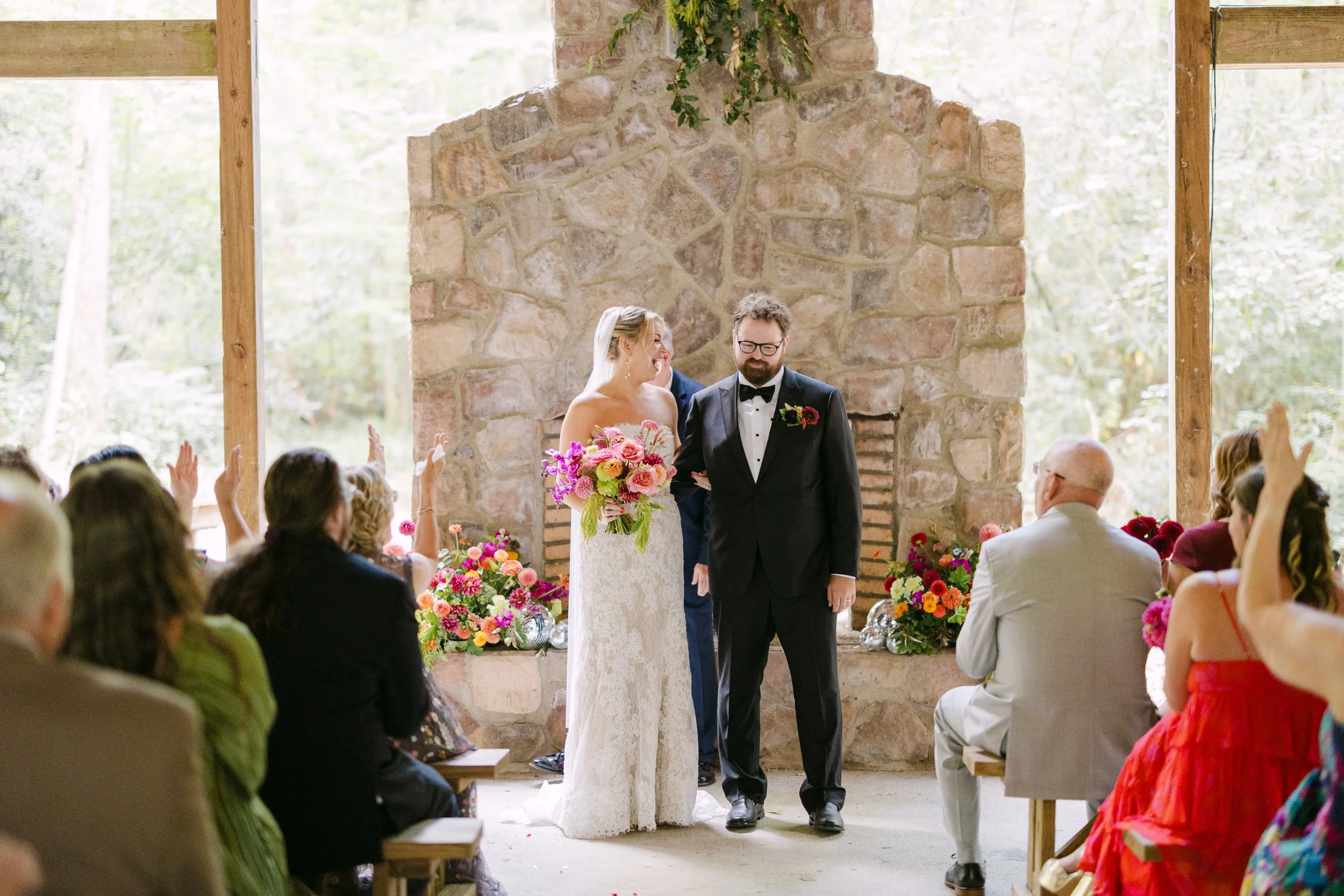 A bride and groom during their wedding ceremony, standing in front of a large stone fireplace with floral decorations, surrounded by seated guests raising their hands.