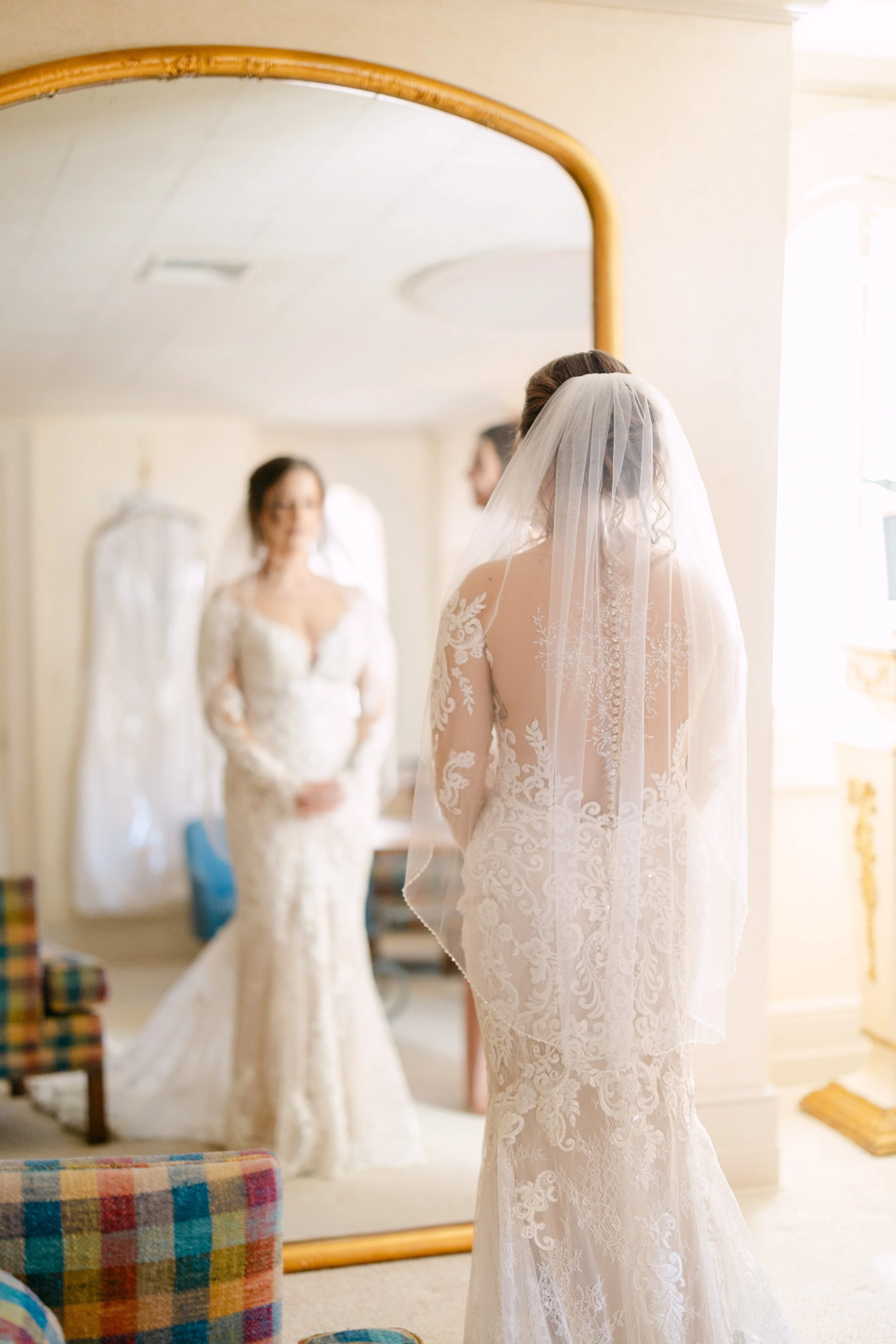 Bride in a lace wedding dress and veil looks at her reflection in a mirror, with her bridesmaid in a similar dress blurred in the background.