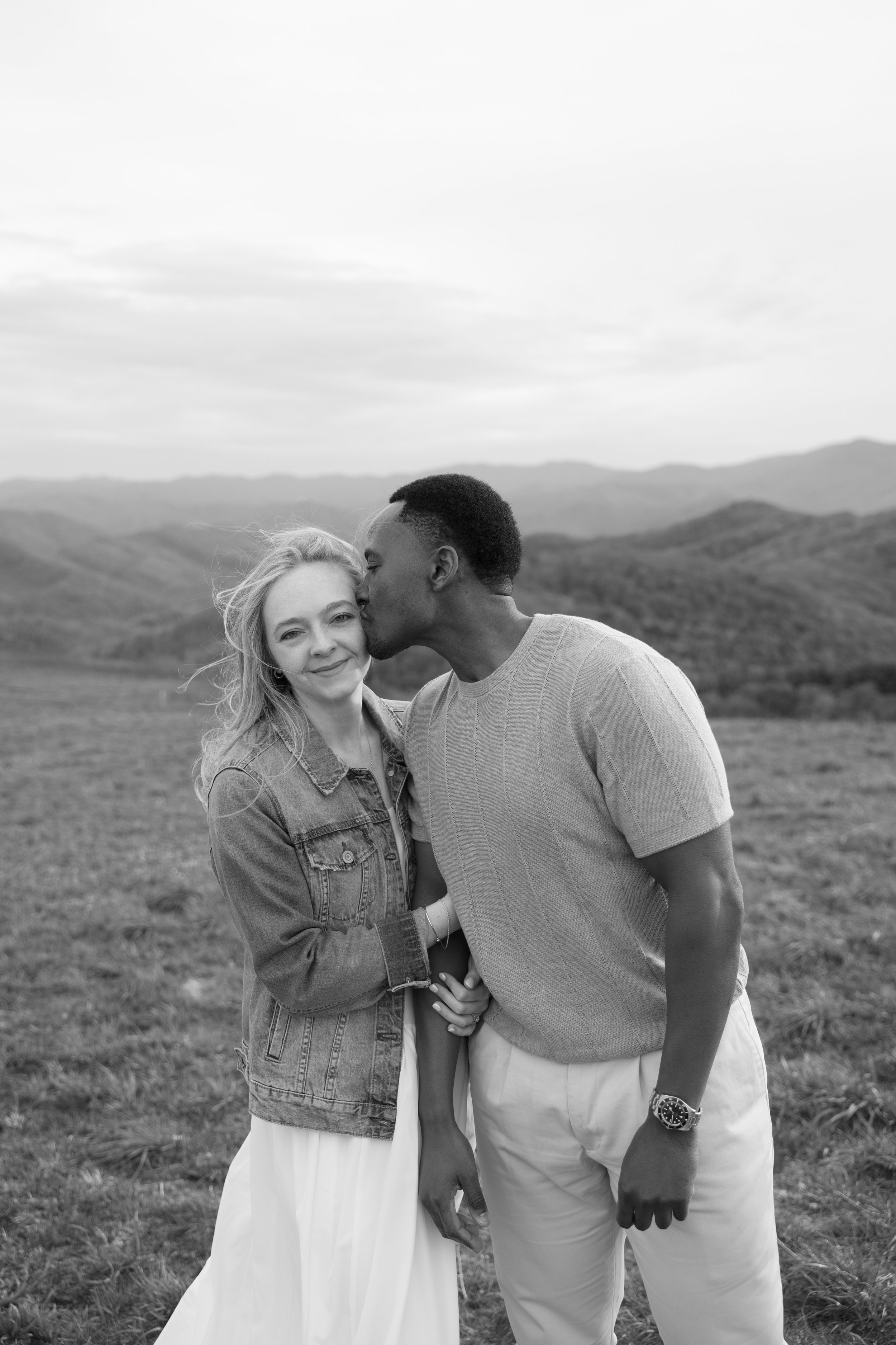 A black and white photo of a couple standing outdoors in a field with mountains in the background. The man is kissing the woman's temple as they hold hands, both smiling softly.