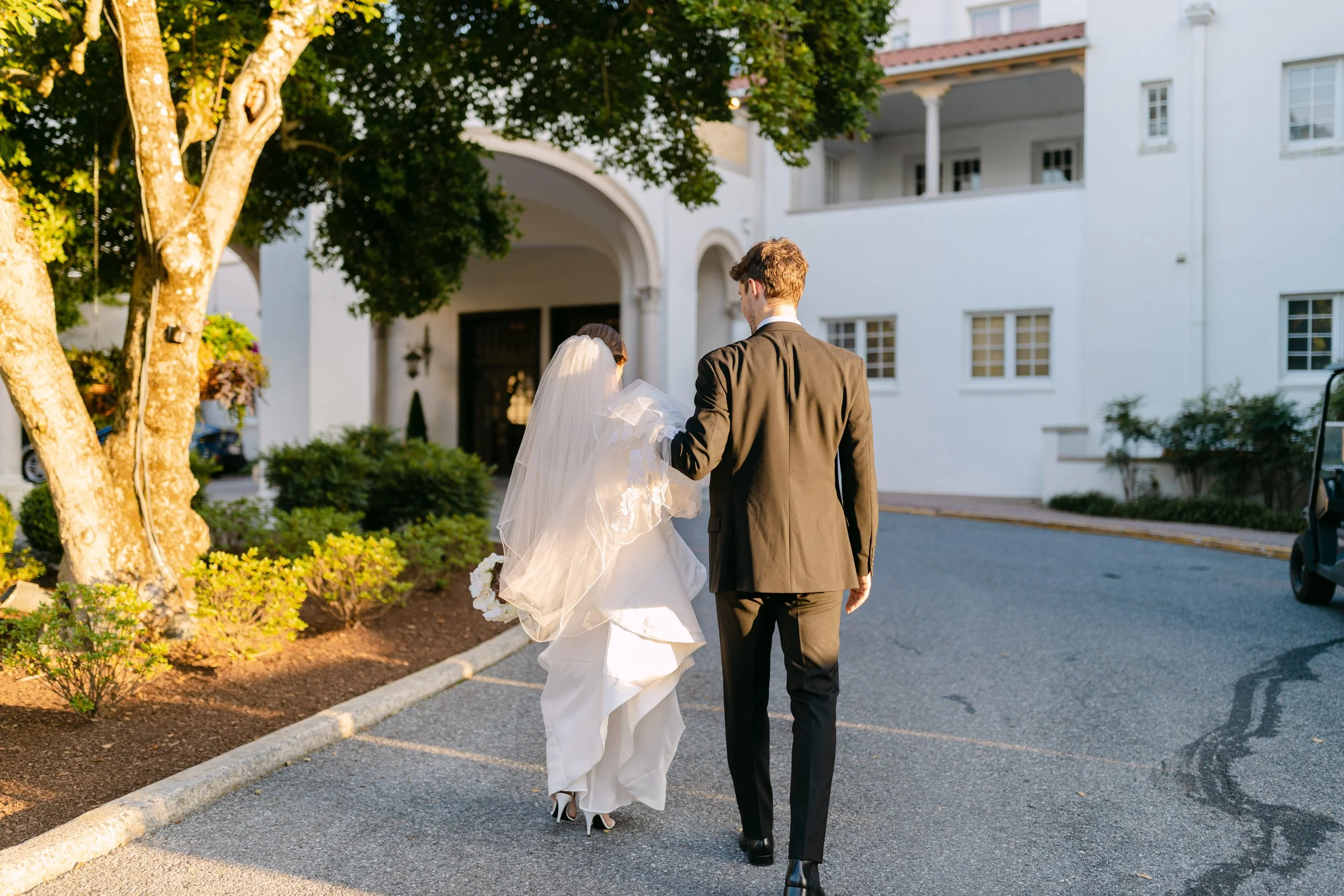 A newlywed couple walking outside on a sunny day. The bride wears a white wedding dress with a veil, and the groom is in a black suit. They are walking towards a white building with greenery around.