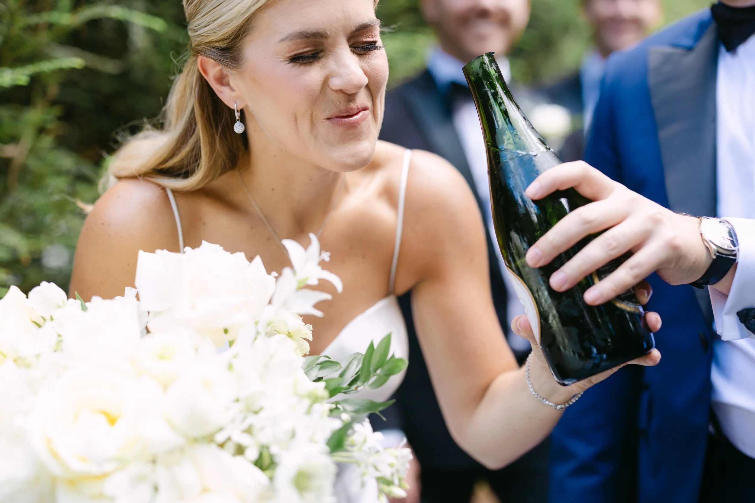 A woman in a wedding dress is holding a flower bouquet and smiling as a person in a suit offers her a bottle of beer, with two men in suits in the background during an outdoor celebration.