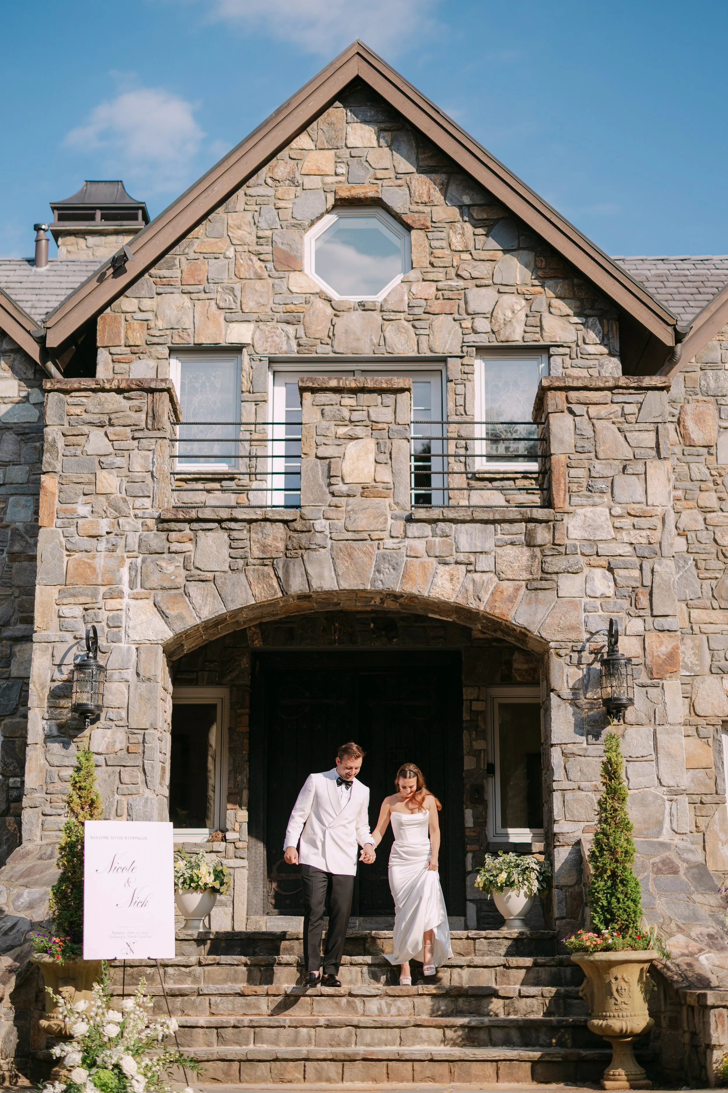 A newlywed couple is walking down stone steps outside a stone house with a tall arched entryway, two small potted plants on either side of the steps, and a wedding sign on the left. The groom is wearing a white tuxedo jacket with a black bow tie, and
