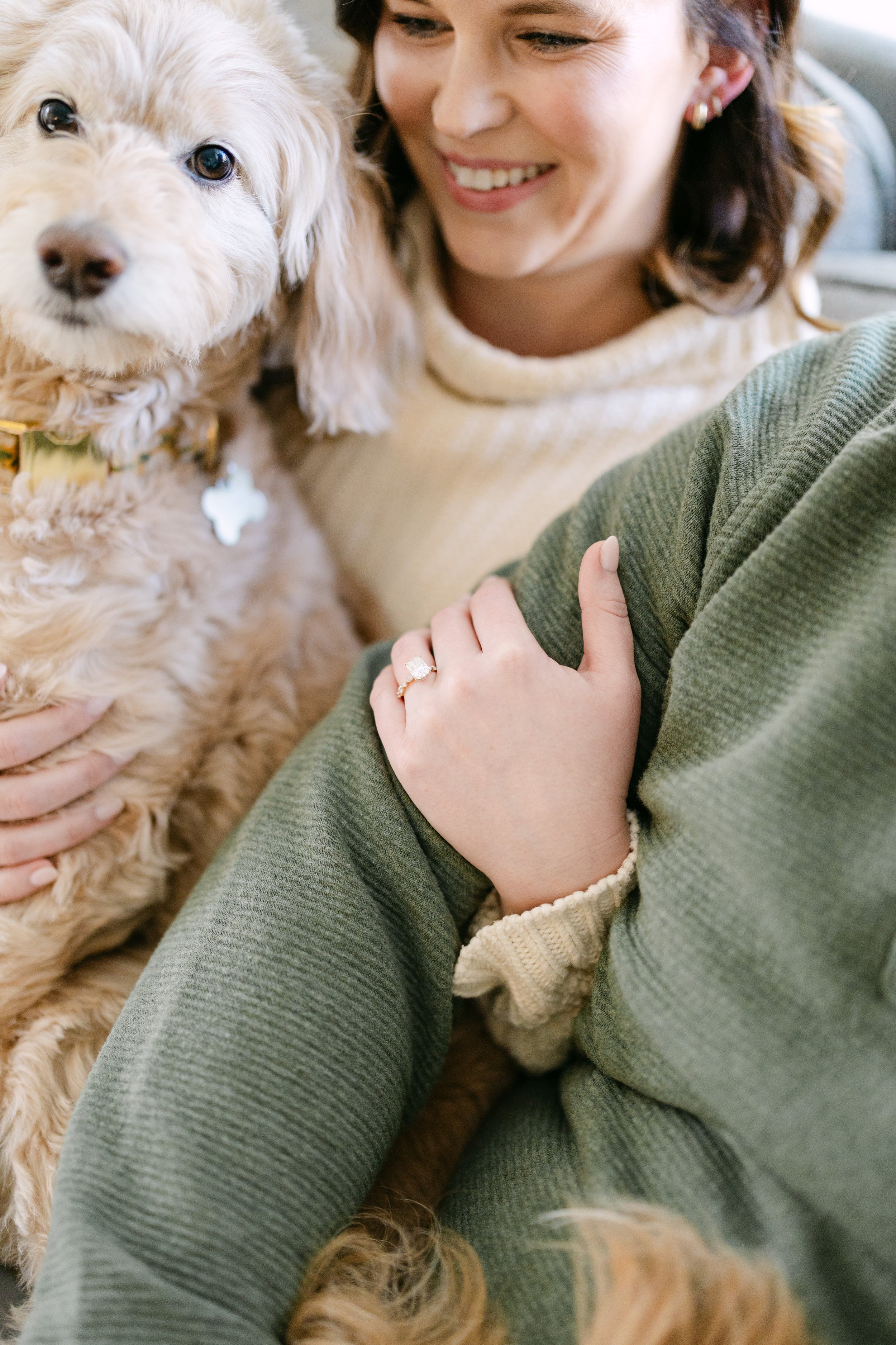 A woman smiling while holding a beige dog, showing her hand with a ring, in a cozy indoor setting.