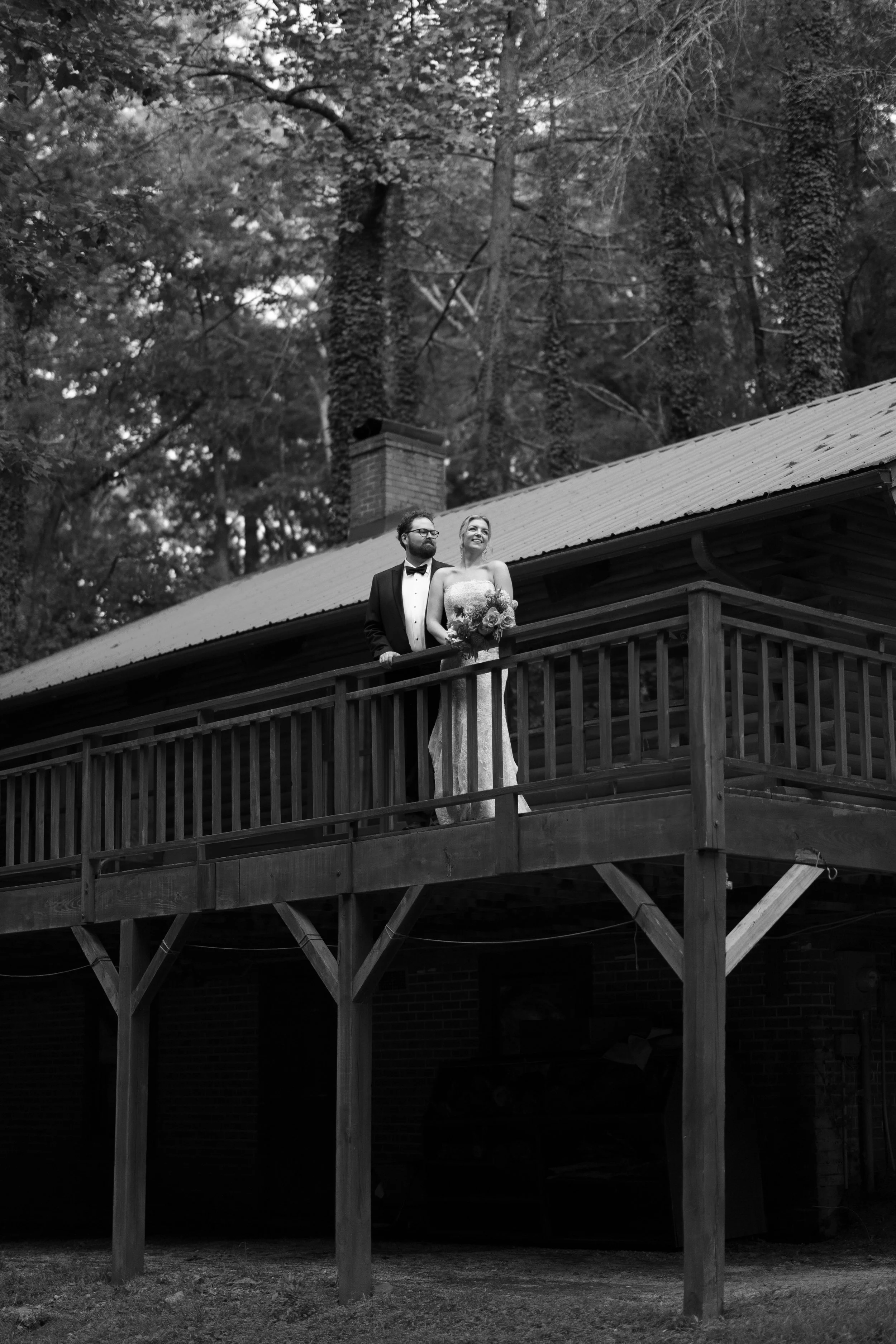 A black and white photo of a bride and groom standing on a wooden balcony outside a house, surrounded by tall trees.