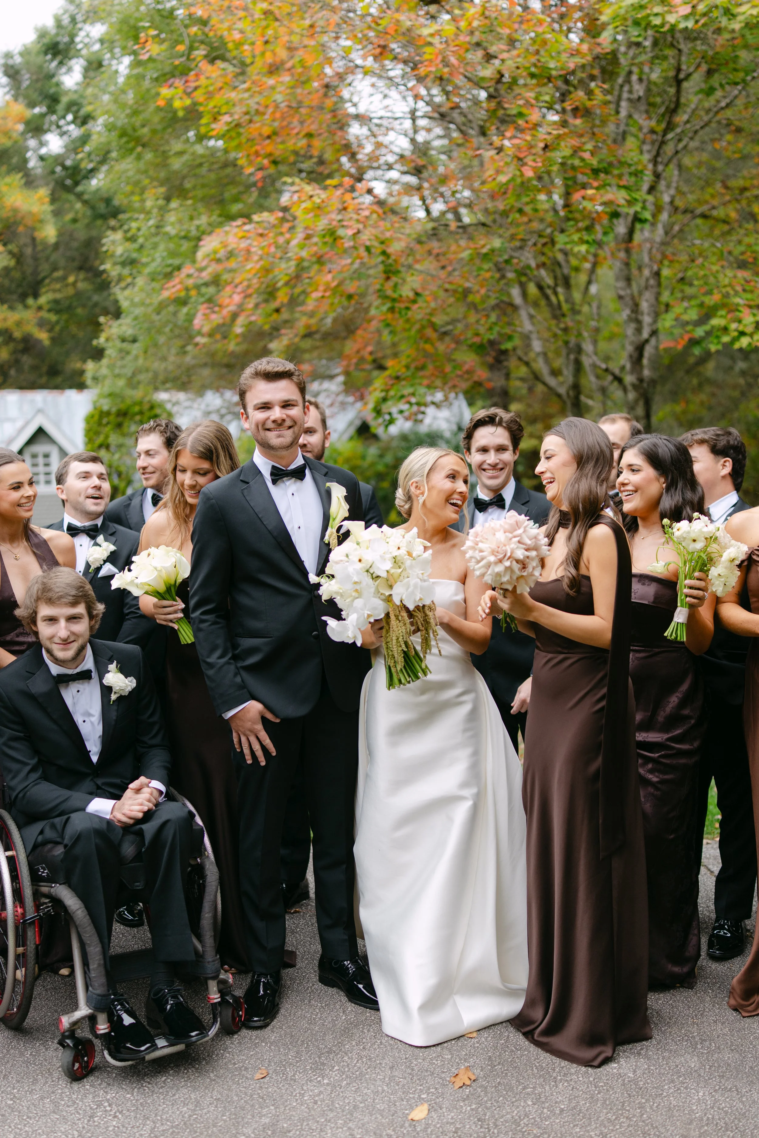 Bride and groom with bridesmaids and groomsmen posing outdoors among autumn trees, holding wedding bouquets.