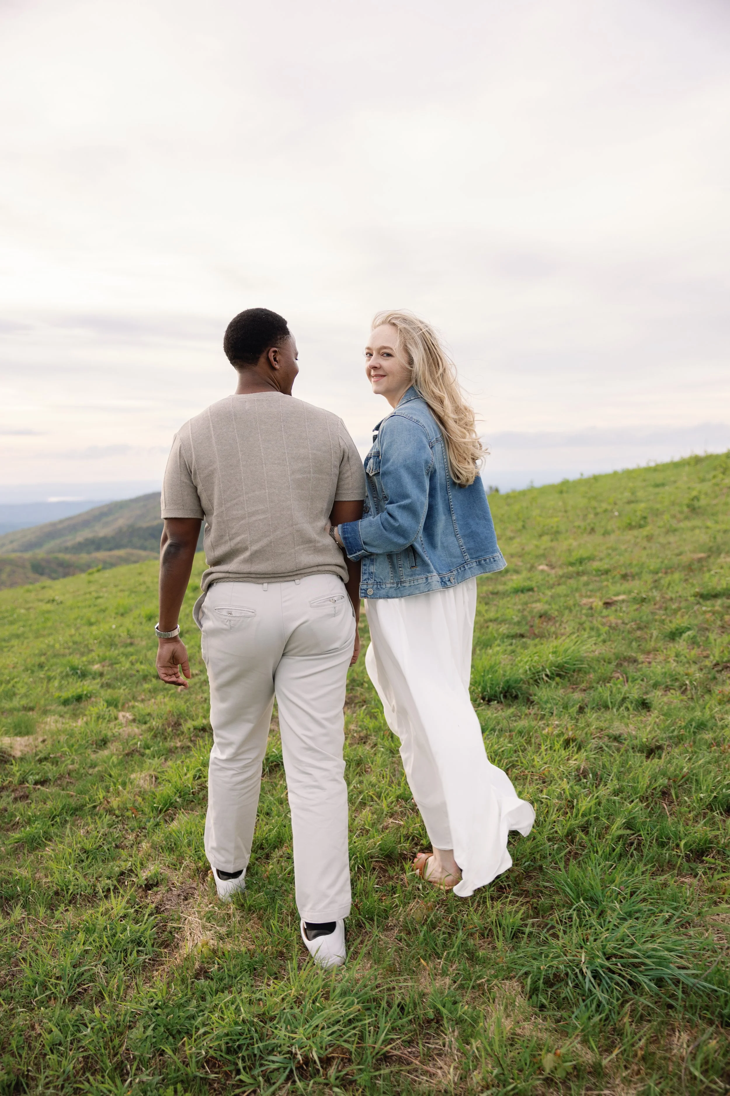 A young man and woman standing in a green field with rolling hills in the background, smiling and looking at each other on a cloudy day.