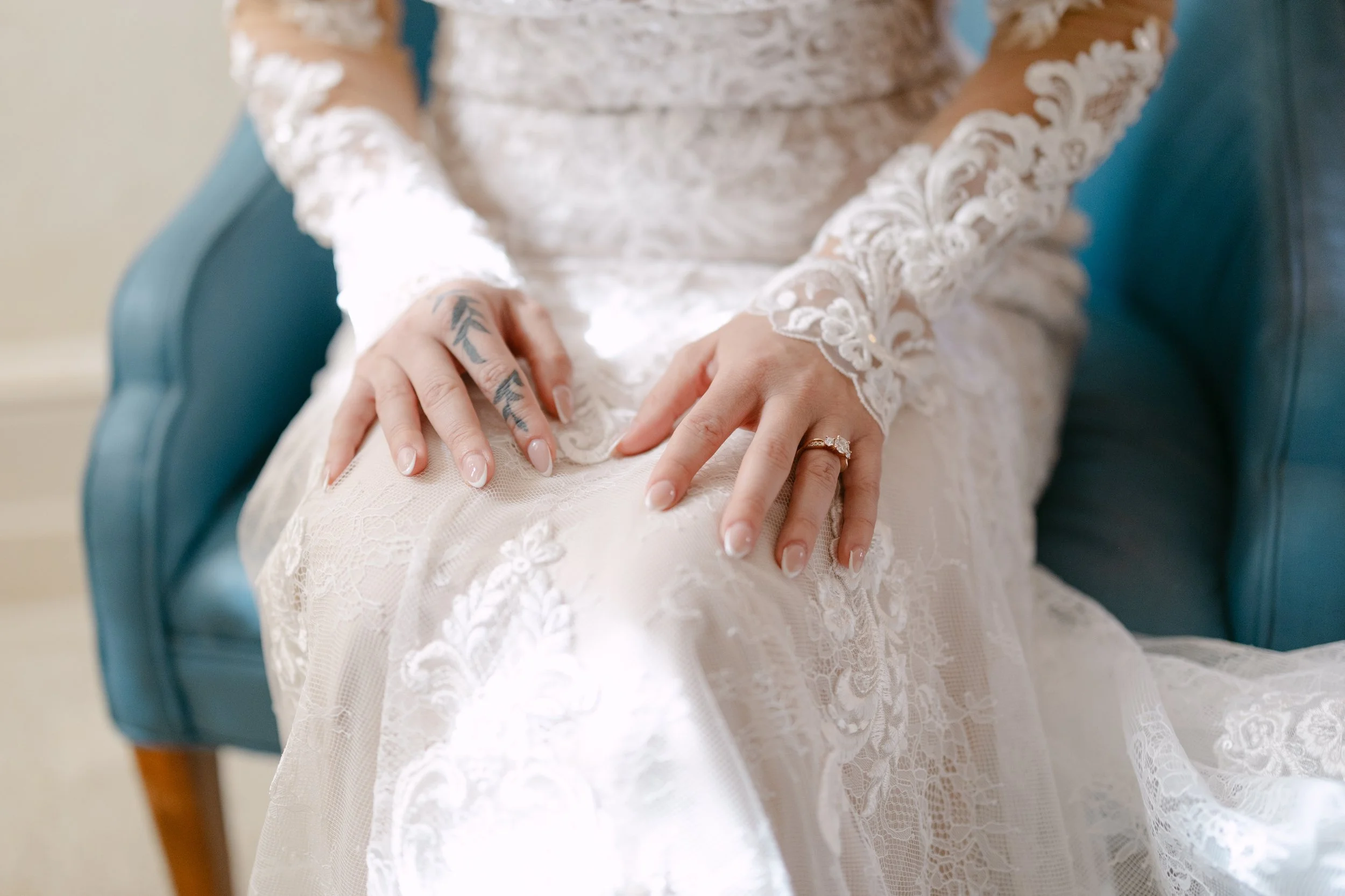 A bride sitting on a blue chair, wearing a lace wedding dress, showing her hands with a wedding ring and tattoos on her fingers.