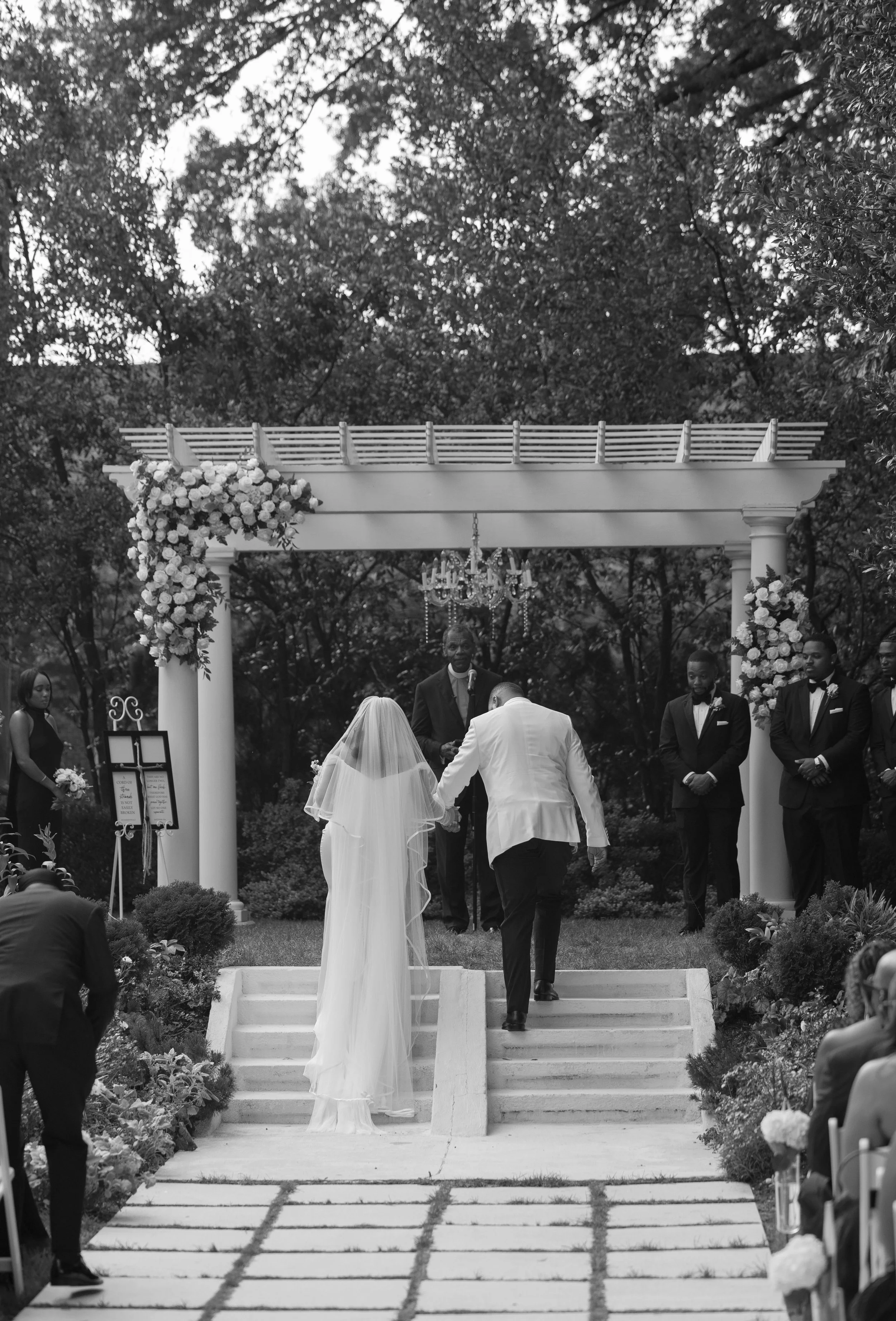 A black and white photo of a wedding ceremony taking place outdoors under a decorated archway with flowers, with the bride and groom holding hands and walking up the steps as the officiant and groomsmen look on, surrounded by guests seated on either 