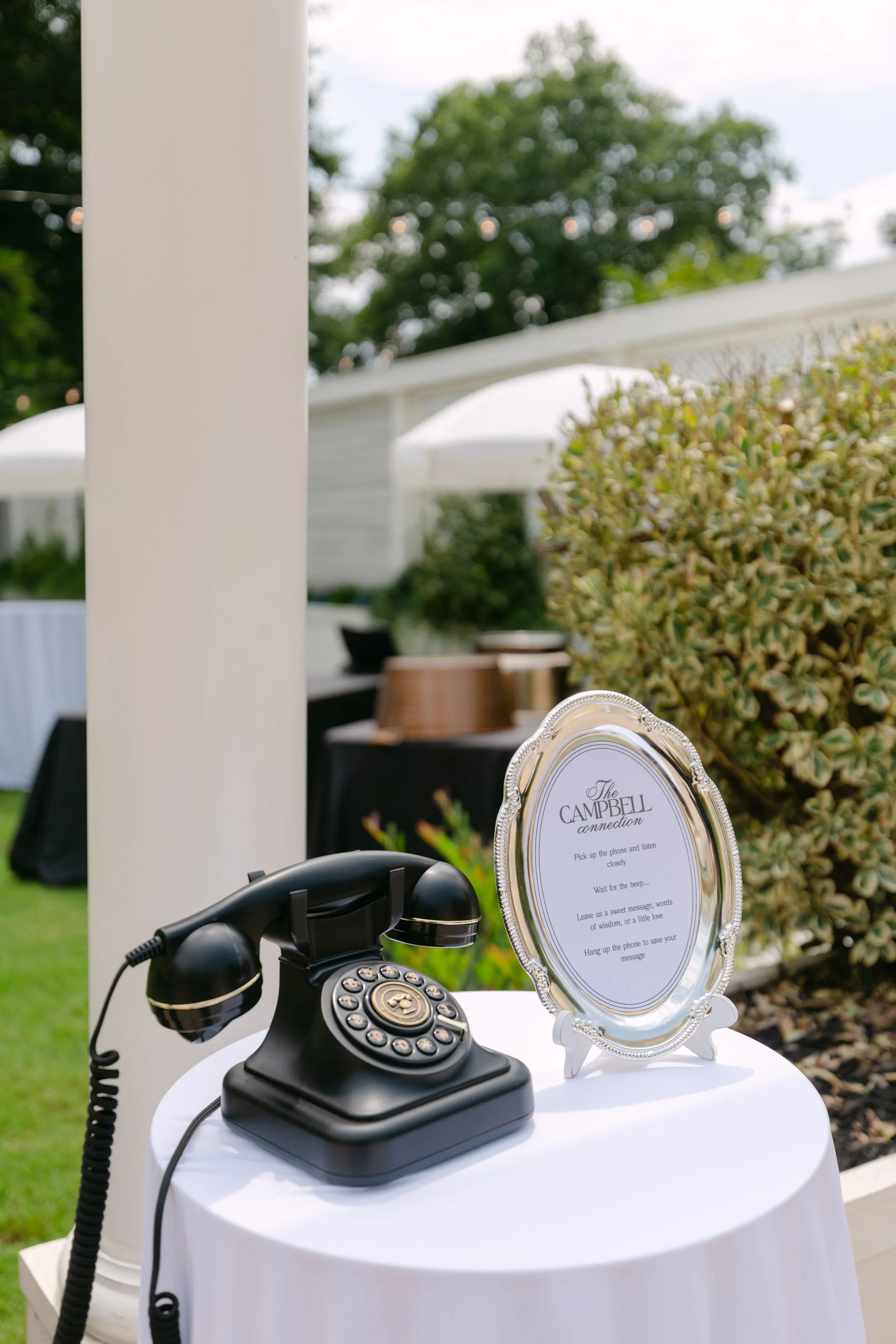 Outdoor setup with a black rotary telephone on a small white table, a decorative frame with signage that reads 'The Campbell connection,' and a plant in a pot, with greenery and outdoor umbrellas in the background.