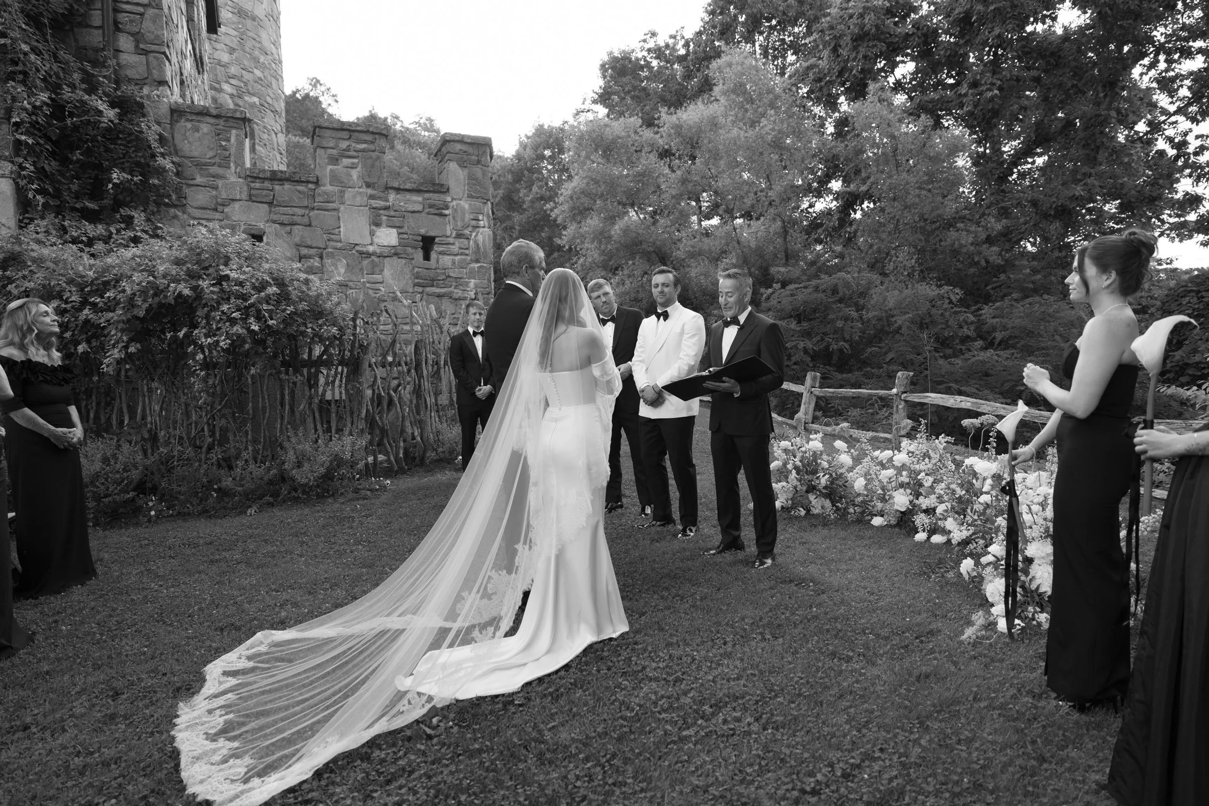 A wedding ceremony outdoors with the bride and groom facing each other, surrounded by bridesmaids and groomsmen. The bridesmaids are in dark dresses, and the groomsmen in tuxedos. The officiant is reading from a book. The scene is set near a stone ca