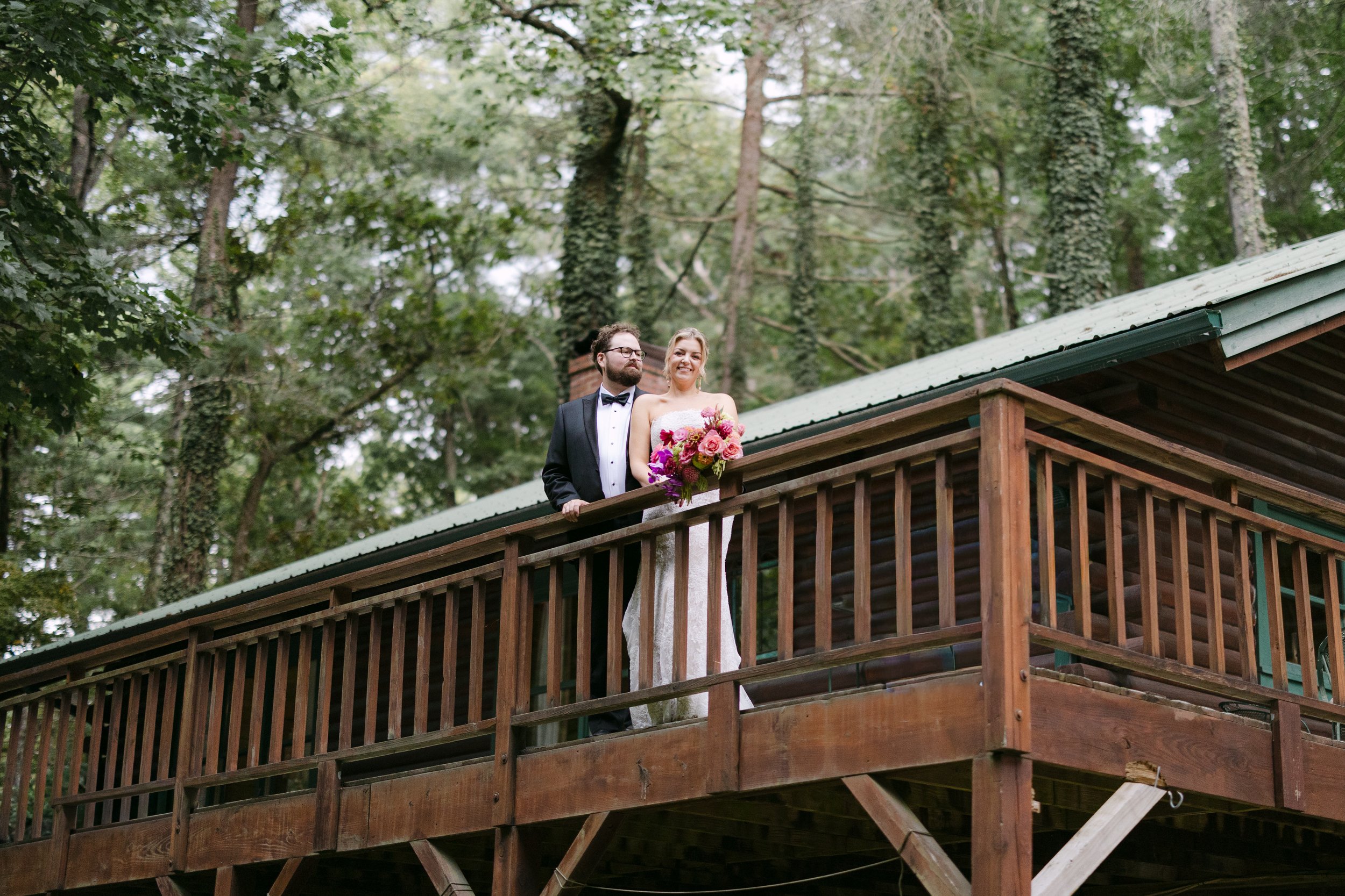 A bride and groom standing on a wooden balcony in a forest, with the bride holding a bouquet of pink and purple flowers and smiling.