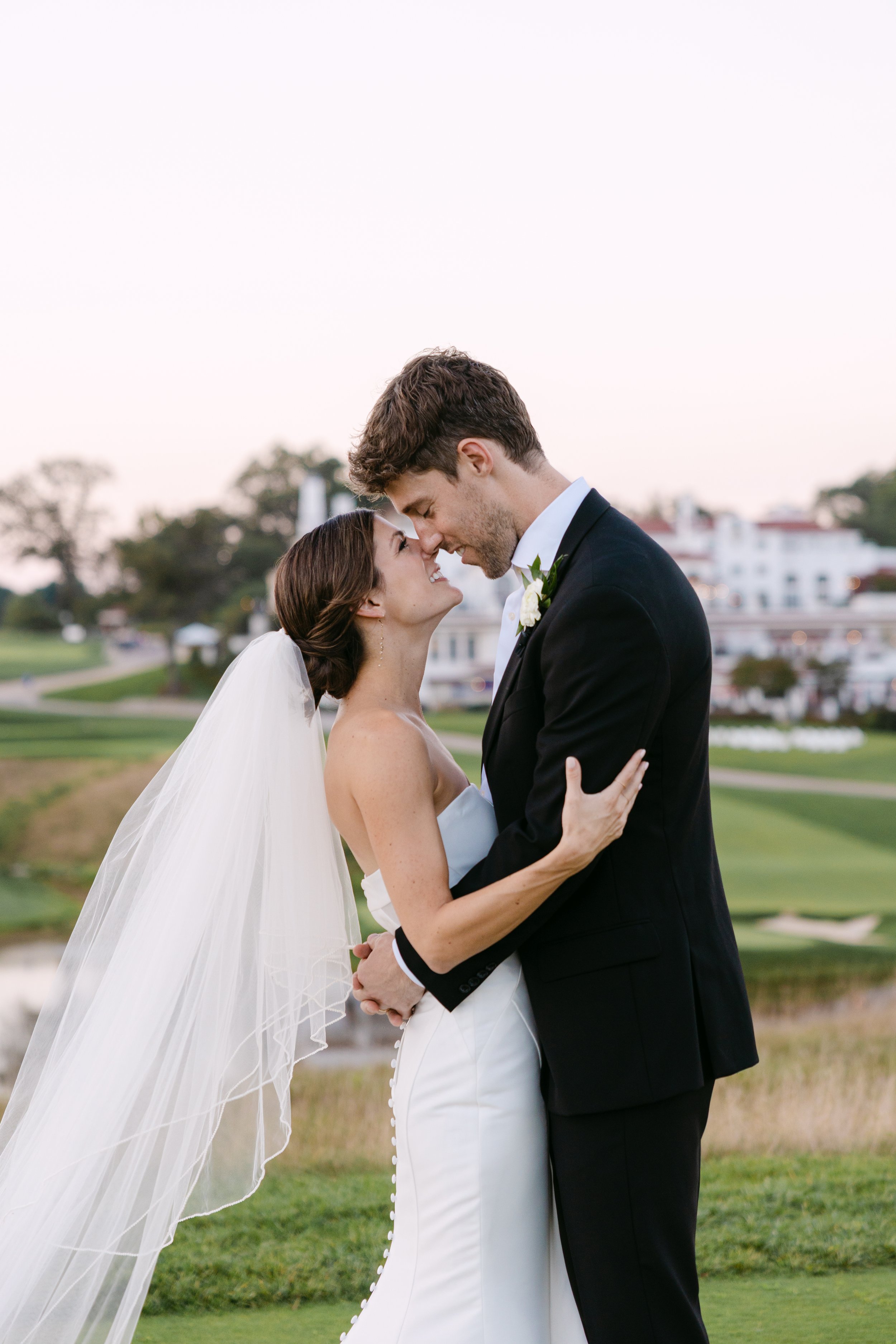 Bride and groom embracing on a golf course during sunset, gazing into each other's eyes
