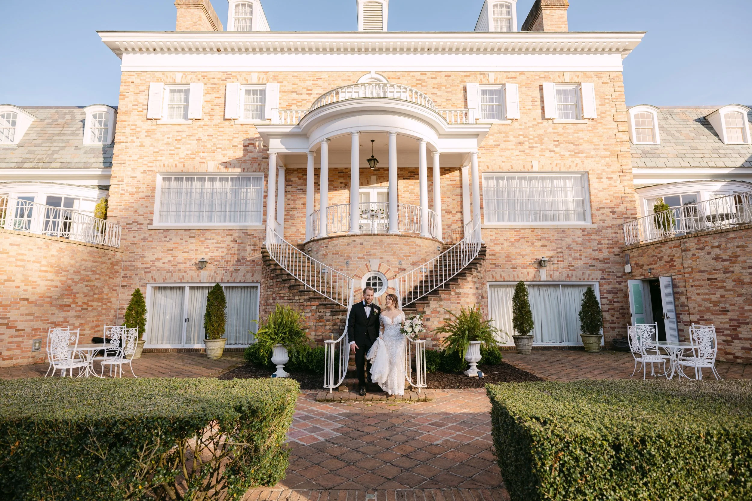 A bride and groom in wedding attire standing in front of a large brick mansion with a curved staircase, windows, and outdoor furniture, celebrating their wedding day.