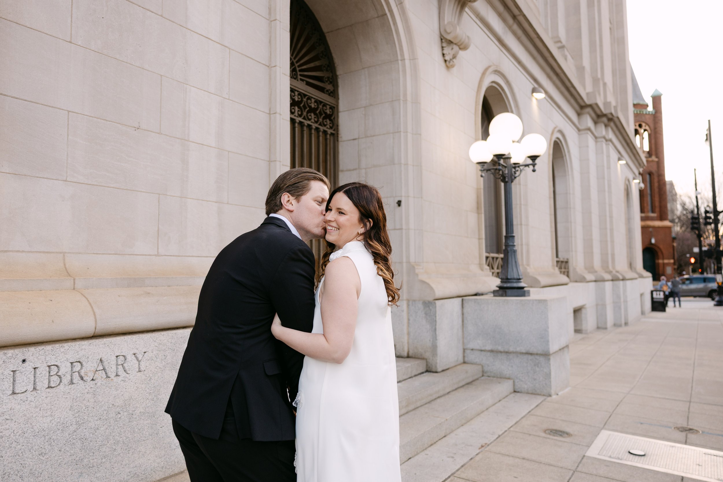 A couple dressed in wedding attire sharing a kiss outdoors near a historic building with large windows, stone walls, and lamp posts.