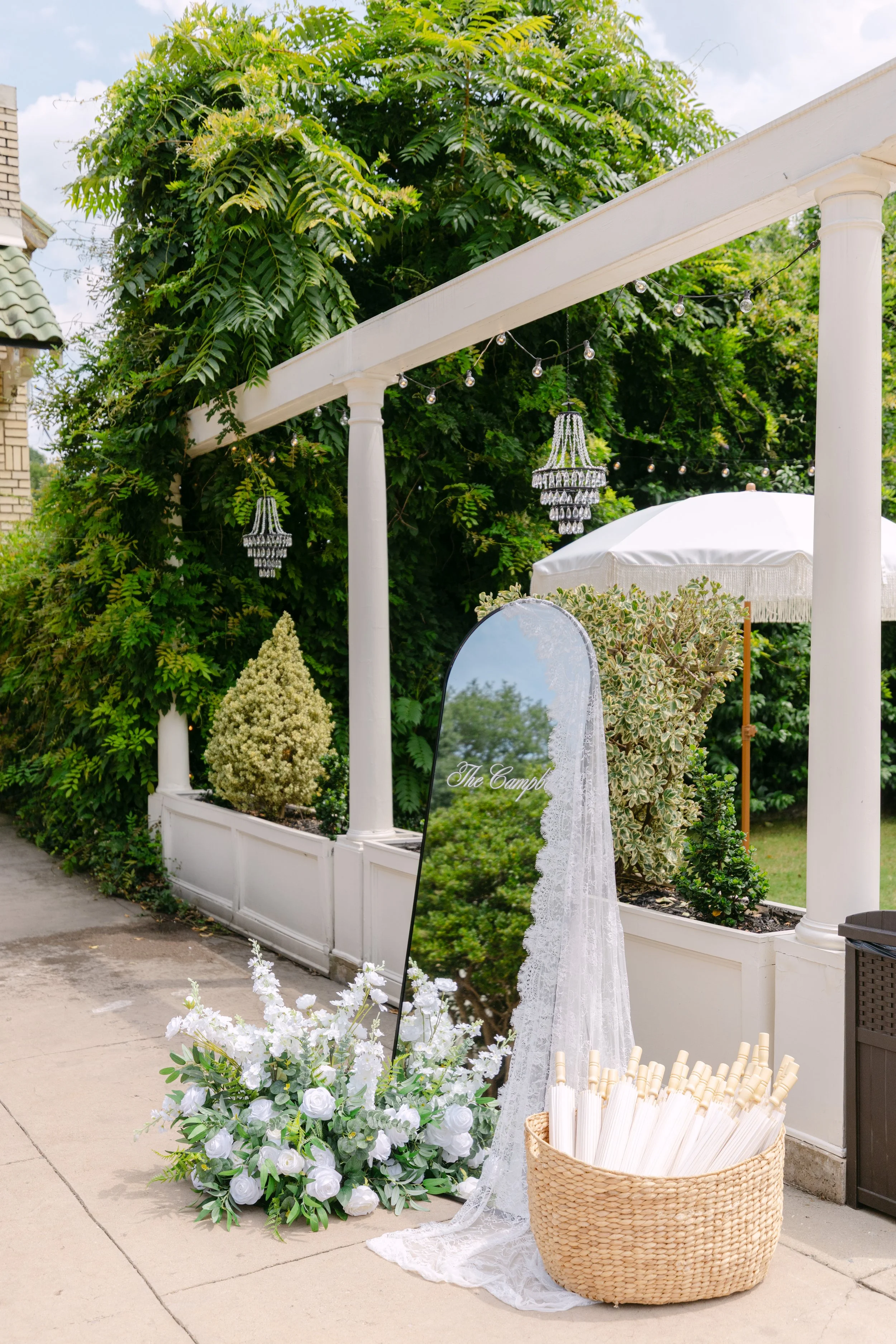 A white floral arch with a sign that reads "The Campbell" and a basket of white umbrellas, with white flowers and greenery in the foreground, decorated for an outdoor wedding or event.