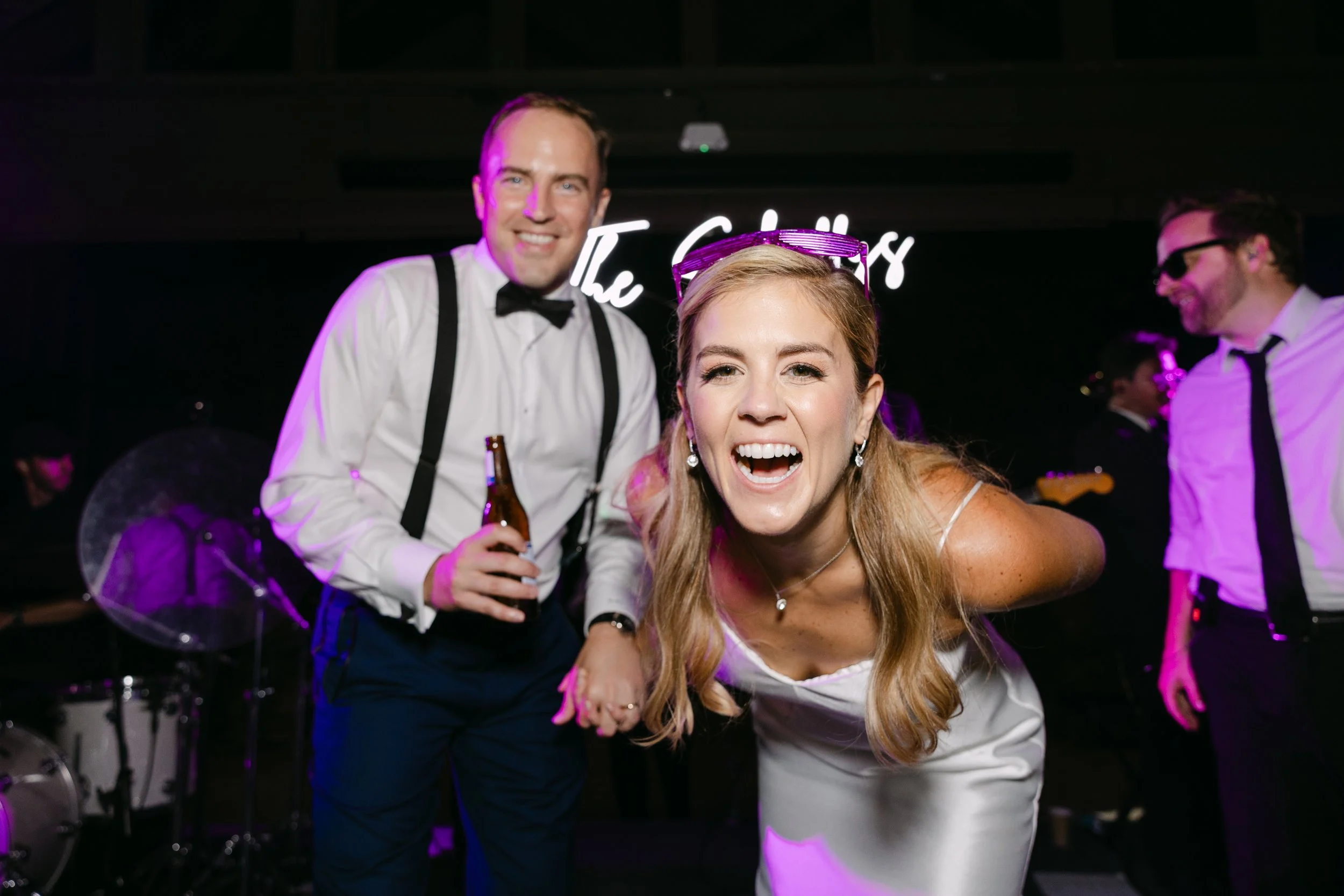 People at a wedding reception, including a woman in a white dress leaning forward and smiling at the camera, a man in a tuxedo holding a beer behind her, and two men in white shirts and black ties in the background, with a band playing on stage and a