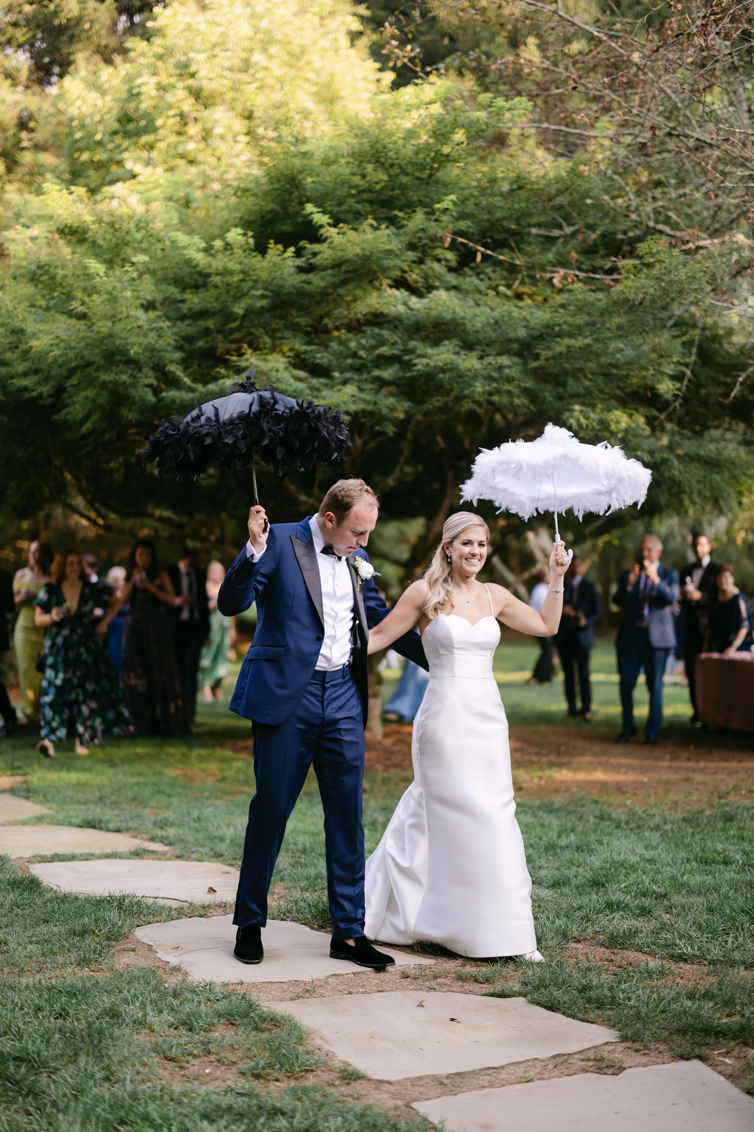 A bride and groom holding black and white umbrellas, walking on a stone path outdoors during their wedding celebration, with guests in the background.