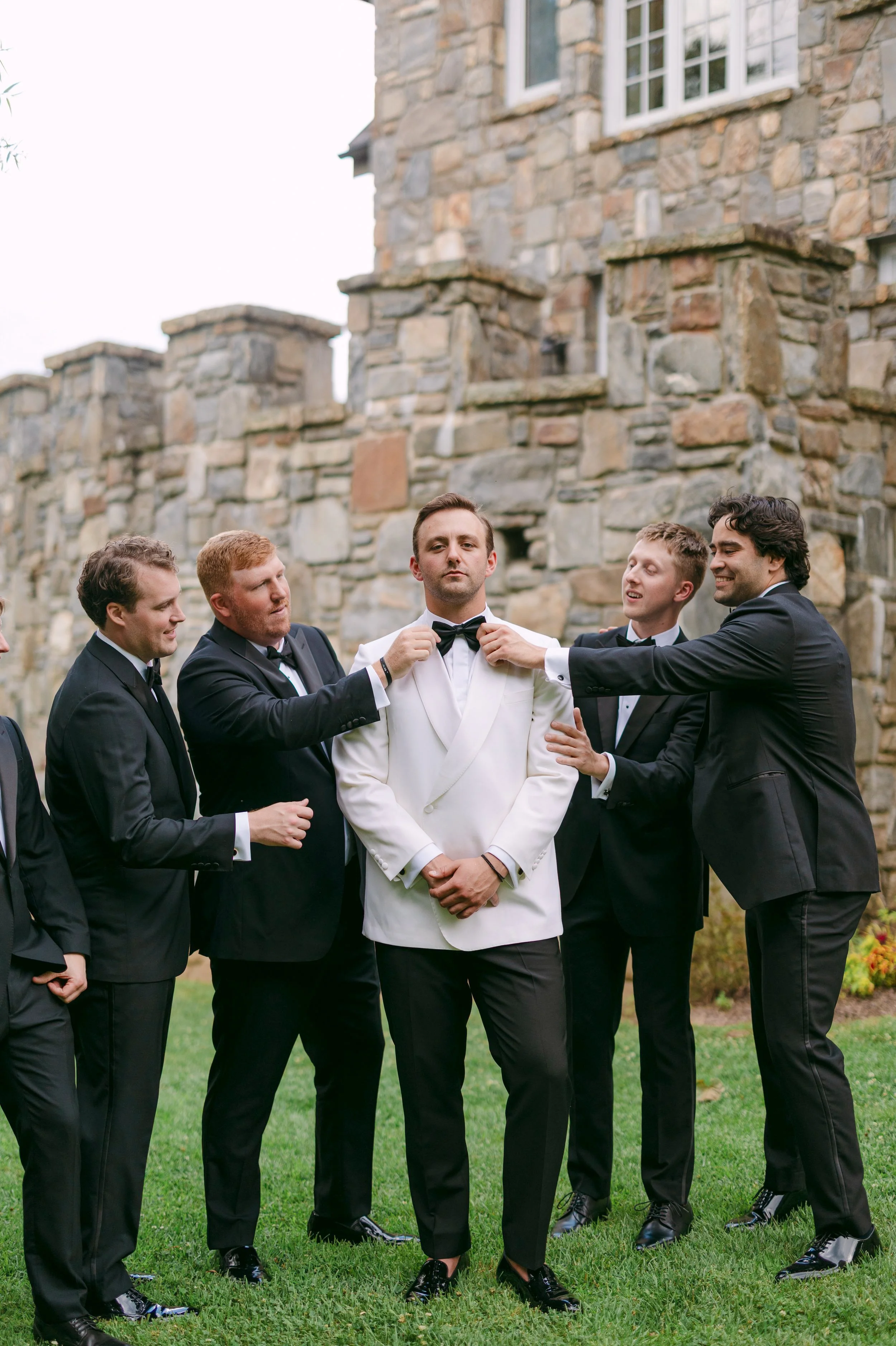 A groom in a white tuxedo stands with friends in tuxedos outdoors, with a stone building in the background, as they adjust his black bow tie.