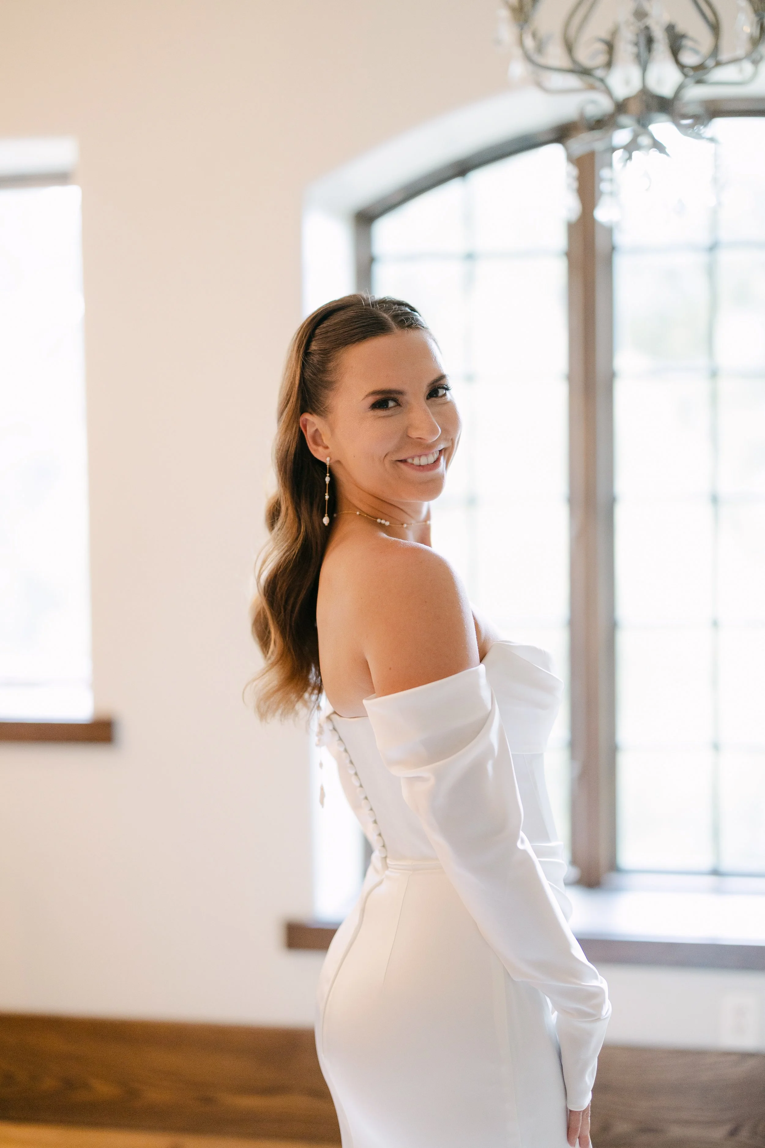 A smiling woman in a white off-shoulder dress with long sleeves, standing near a large window with natural light, indoors with a wooden floor and a chandelier.