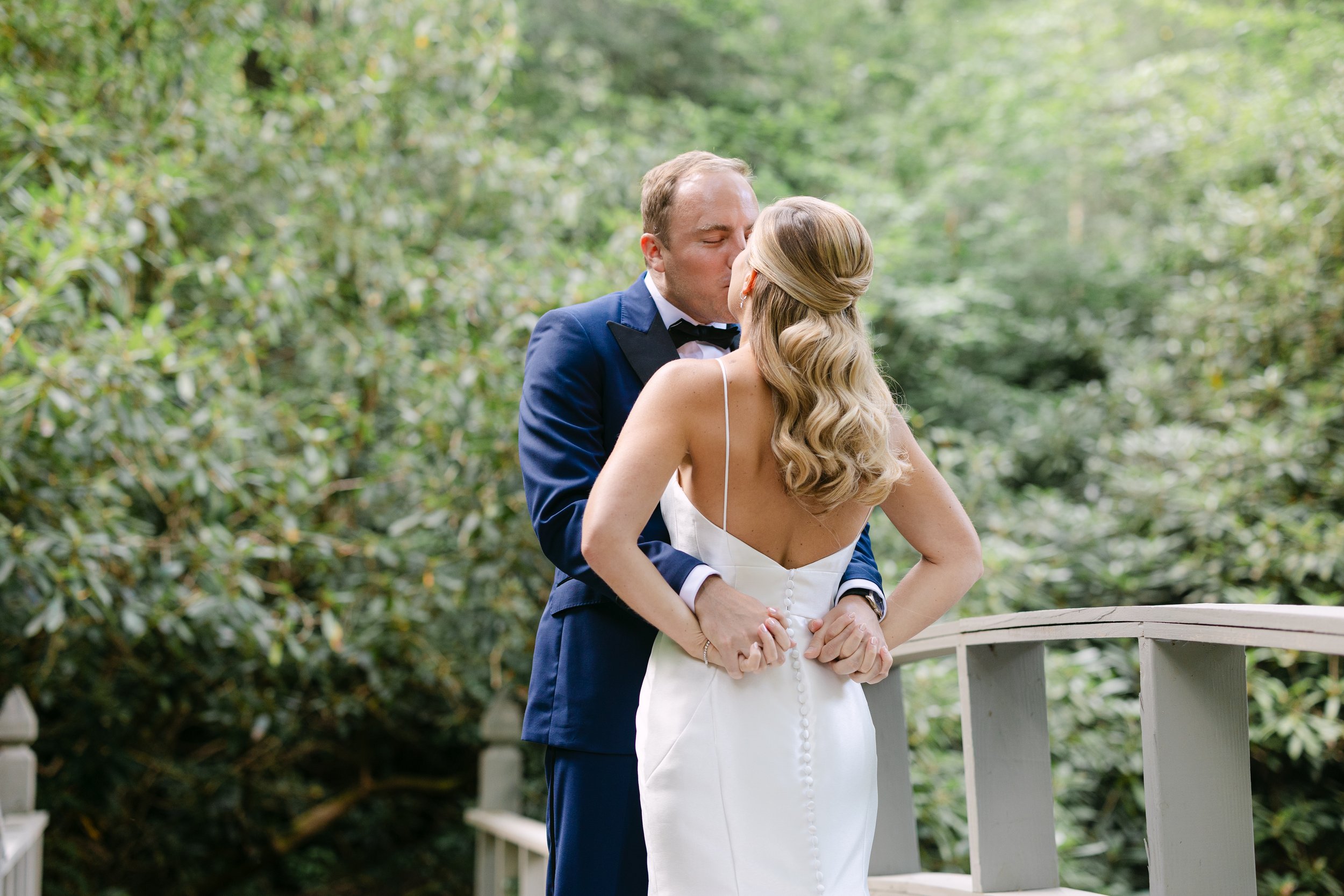 A bride and groom sharing a kiss outdoors surrounded by greenery, with the bride in a white wedding dress and the groom in a navy suit.