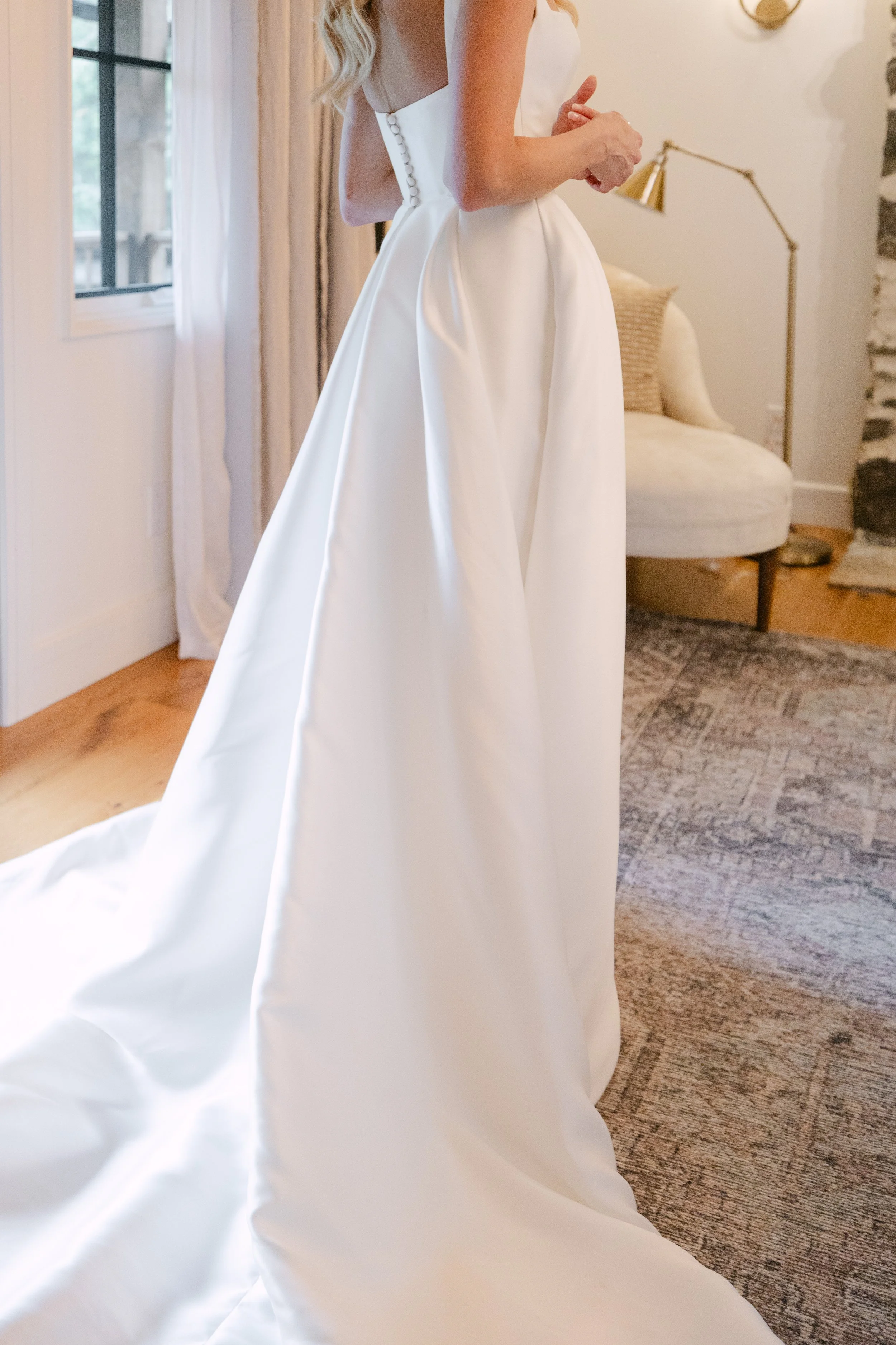 Bride wearing a white wedding dress with a full skirt, standing indoors near a window and a sofa.