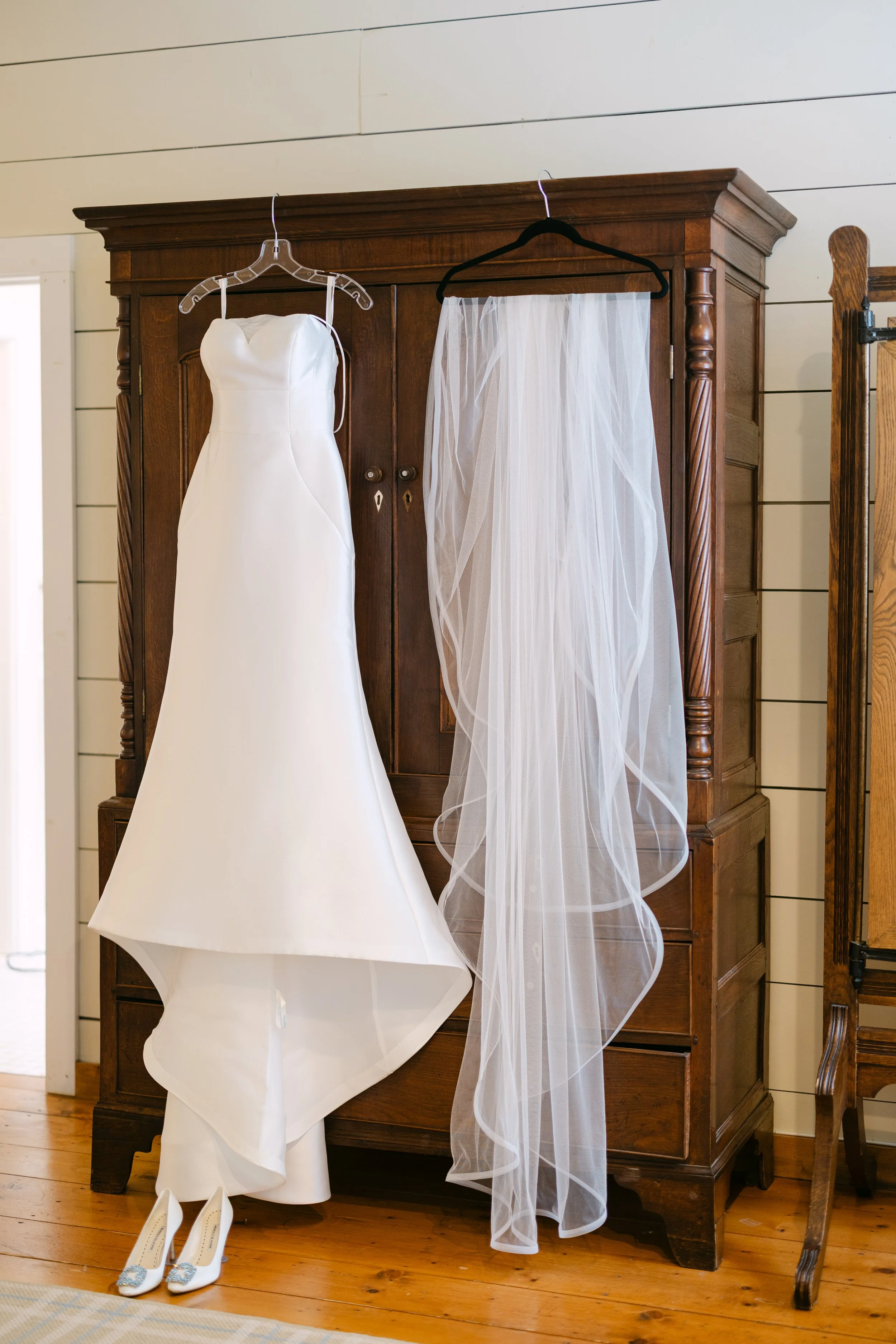 A white wedding dress hanging on a wooden wardrobe, with a veil hanging next to it and a pair of white bridal shoes at the bottom.
