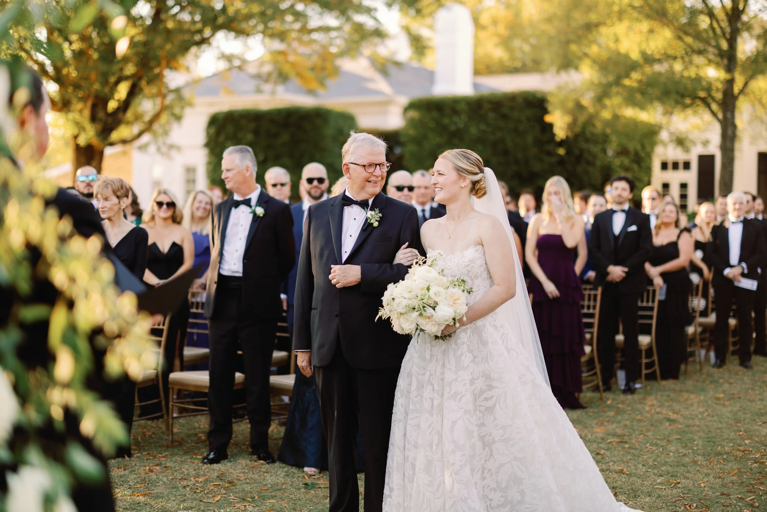 A bride walking down the outdoor wedding aisle with an older man, likely her father, smiling at her, while guests in formal attire stand and watch on a sunny day.