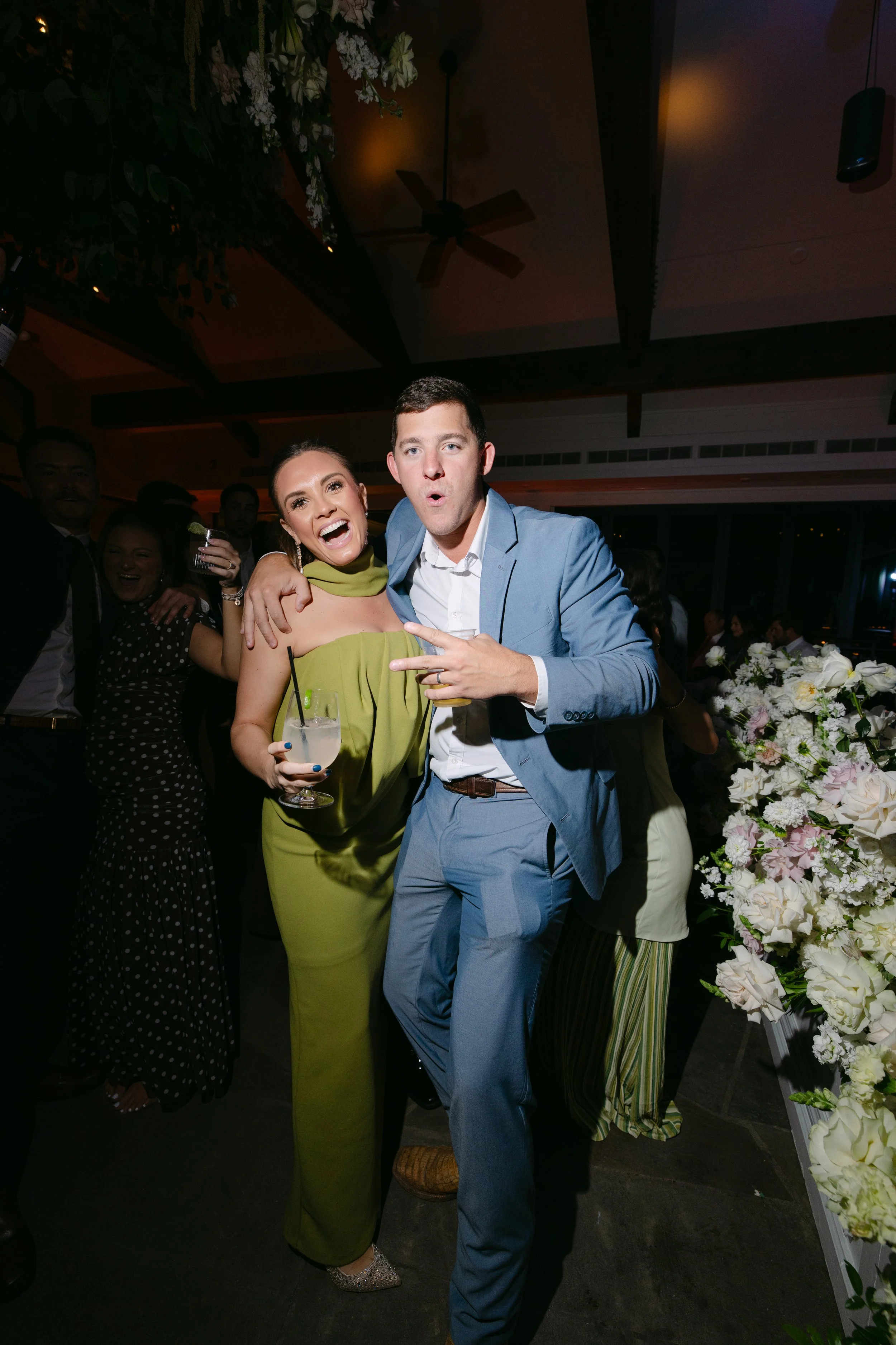Guests smile and pose with drinks at a dimly lit indoor celebration surrounded by floral arrangements.