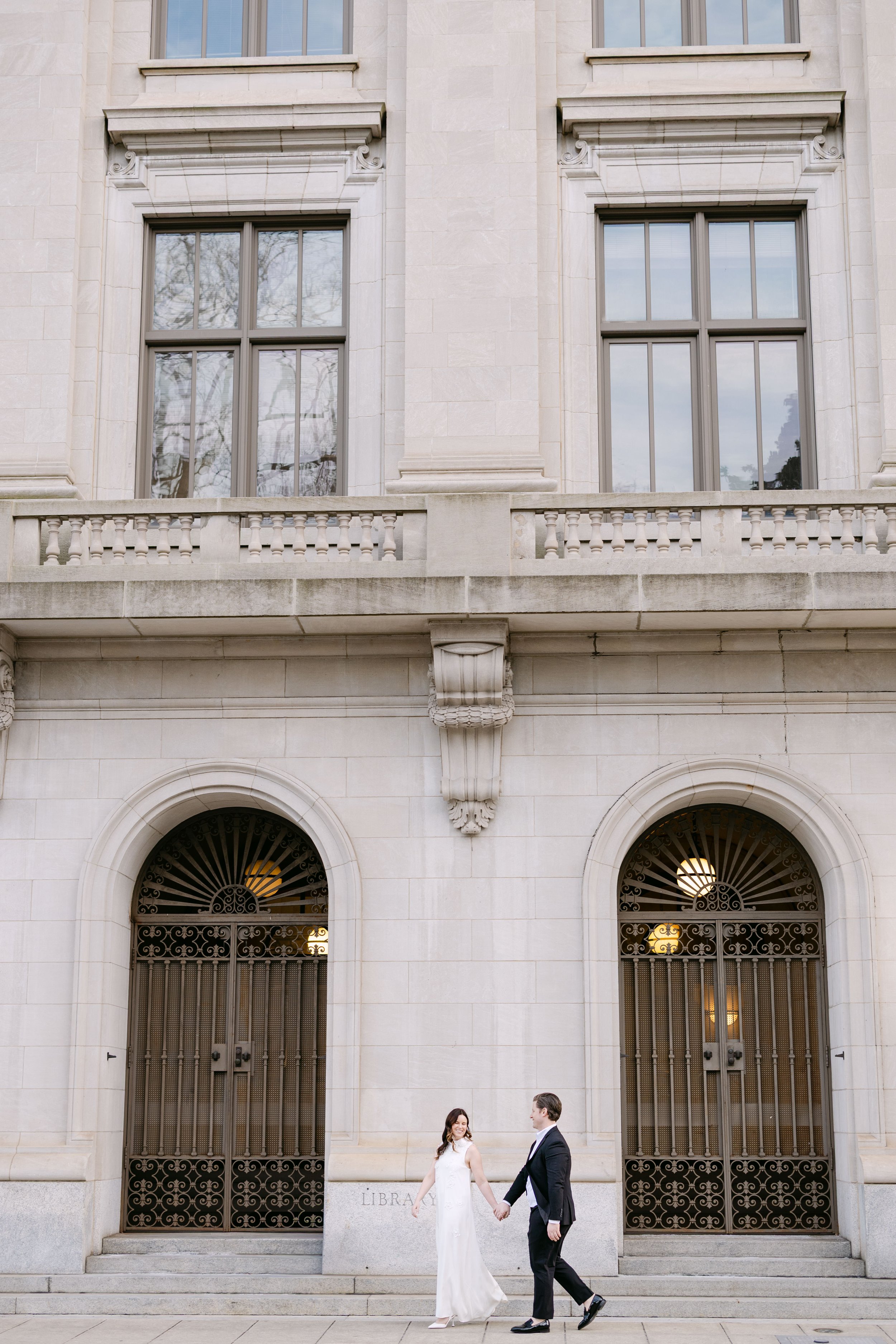 A couple in wedding attire holding hands and walking in front of a large building with arched and barred doors, with the word 'LIBRARY' inscribed near the steps.