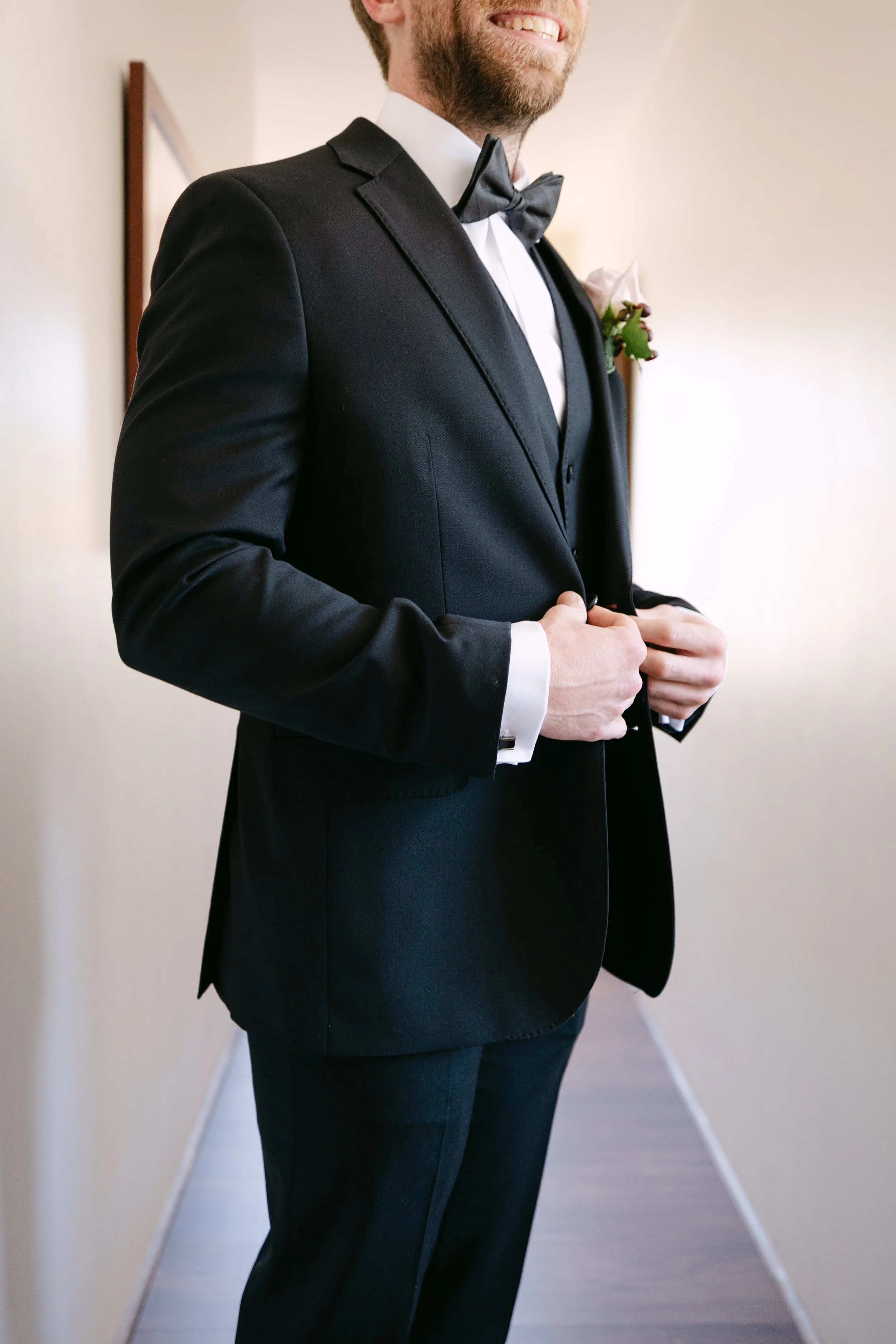 A man dressed in a black tuxedo with a bow tie, adjusting his jacket, standing in a hallway.