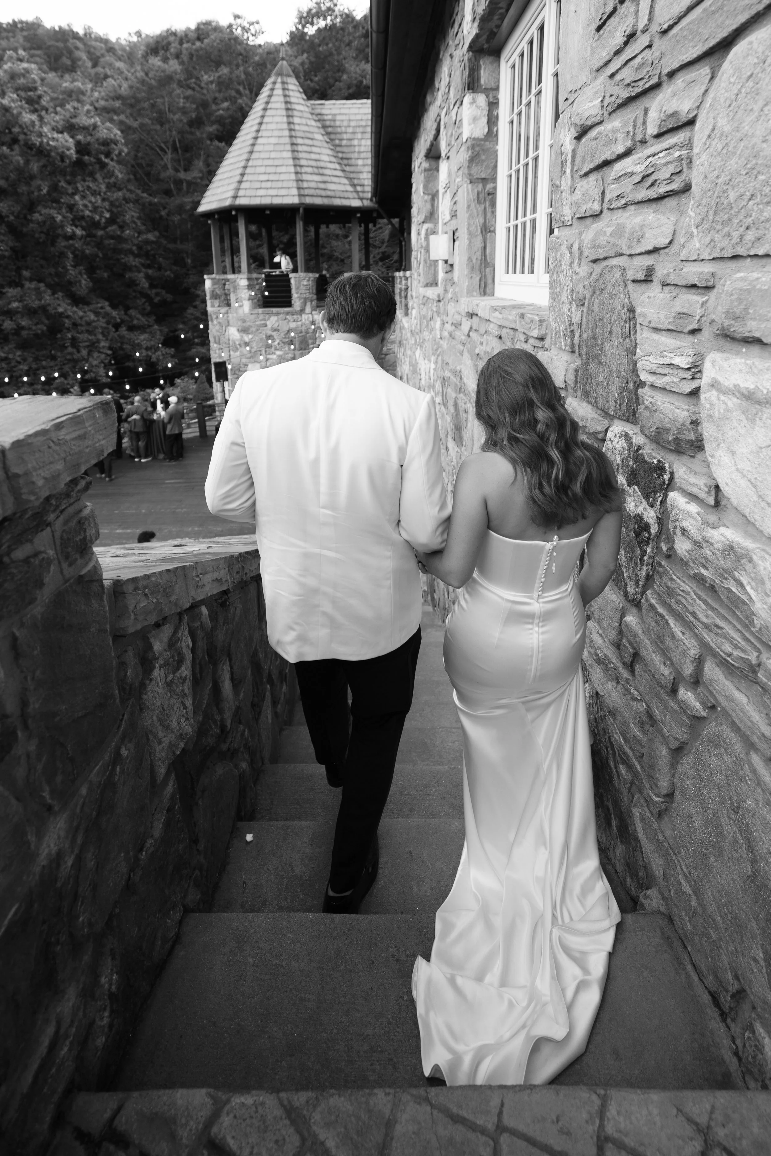 A couple, dressed in formal attire, walking down stone steps at an outdoor event, with a castle-like building and guests in the background.