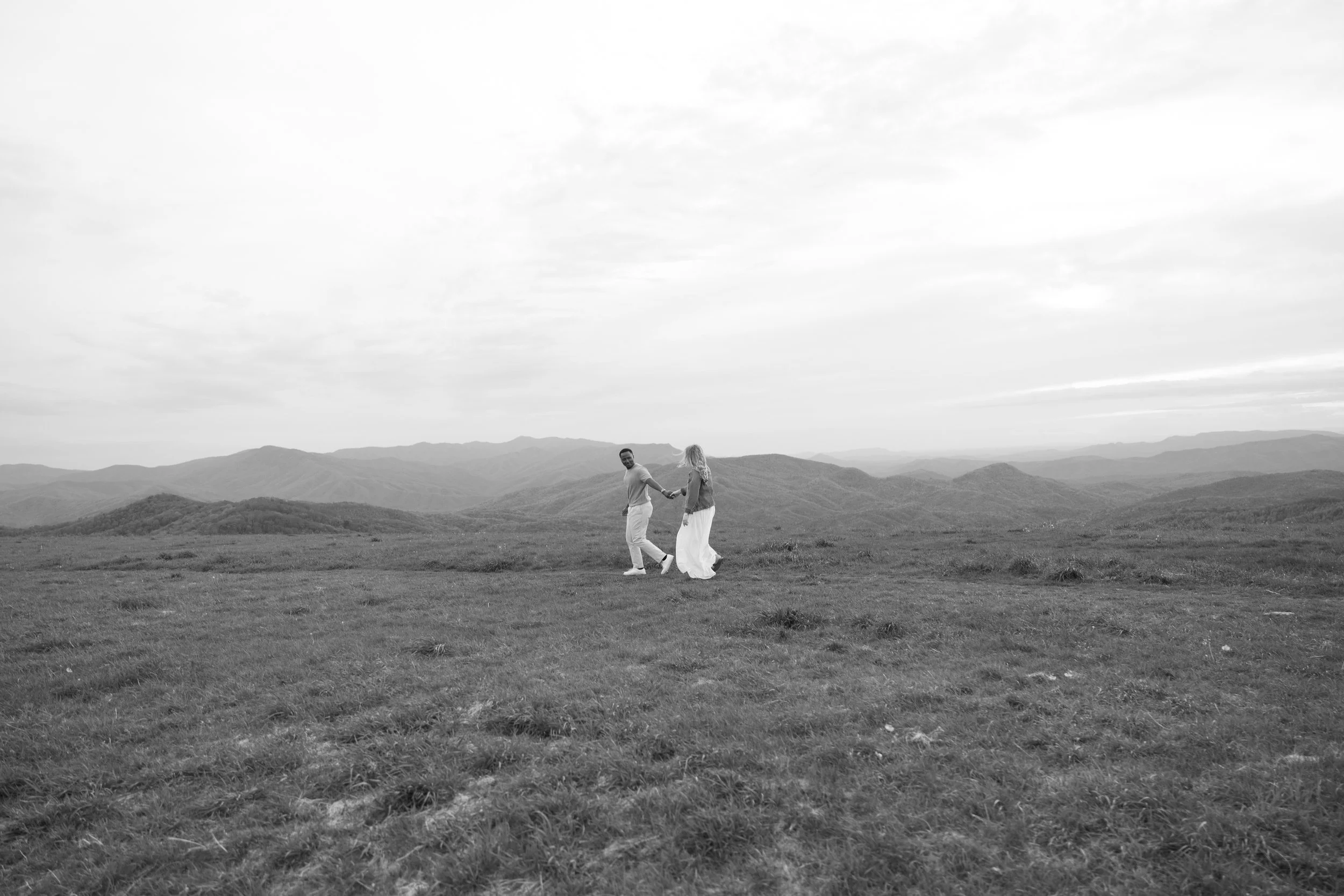 A couple holding hands and walking on a grassy open field with mountain ranges in the background, under a cloudy sky.