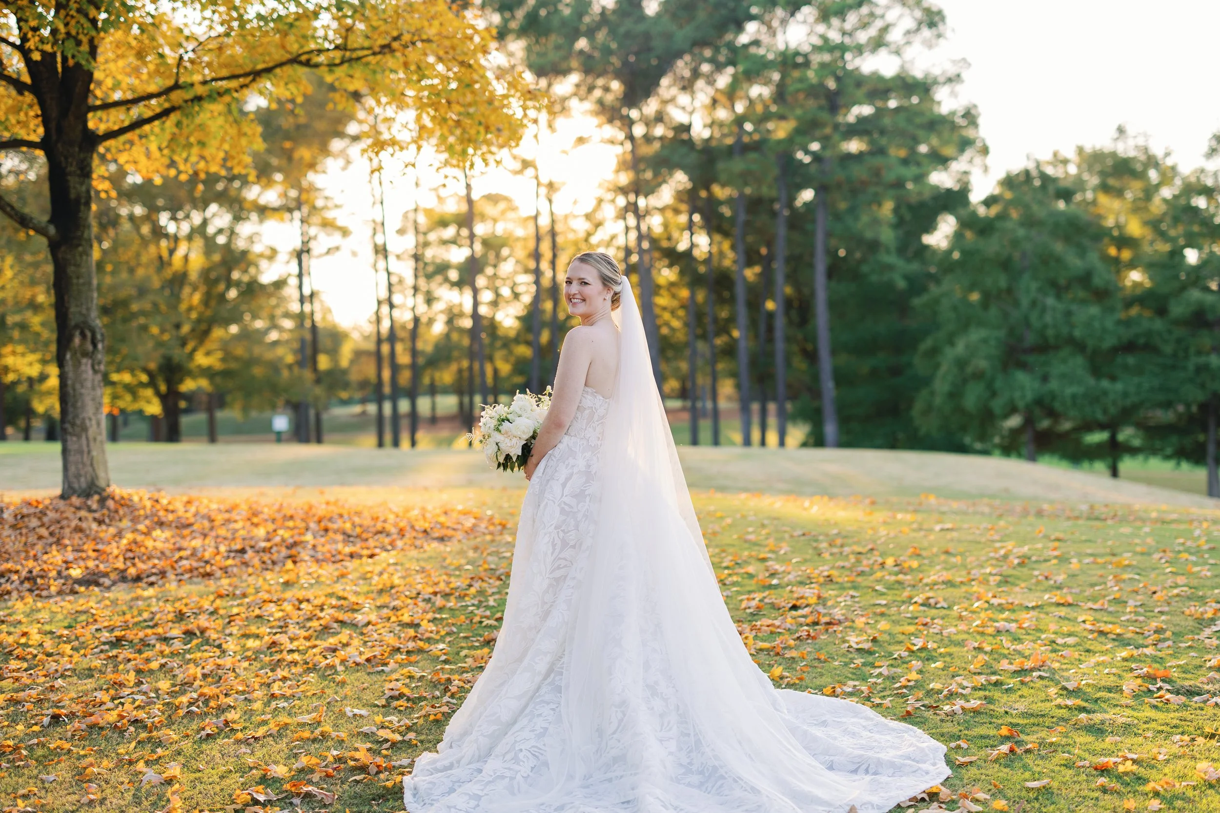 A bride in a white wedding gown holding a bouquet of flowers on a grassy field with autumn leaves, trees, and the setting sun in the background.