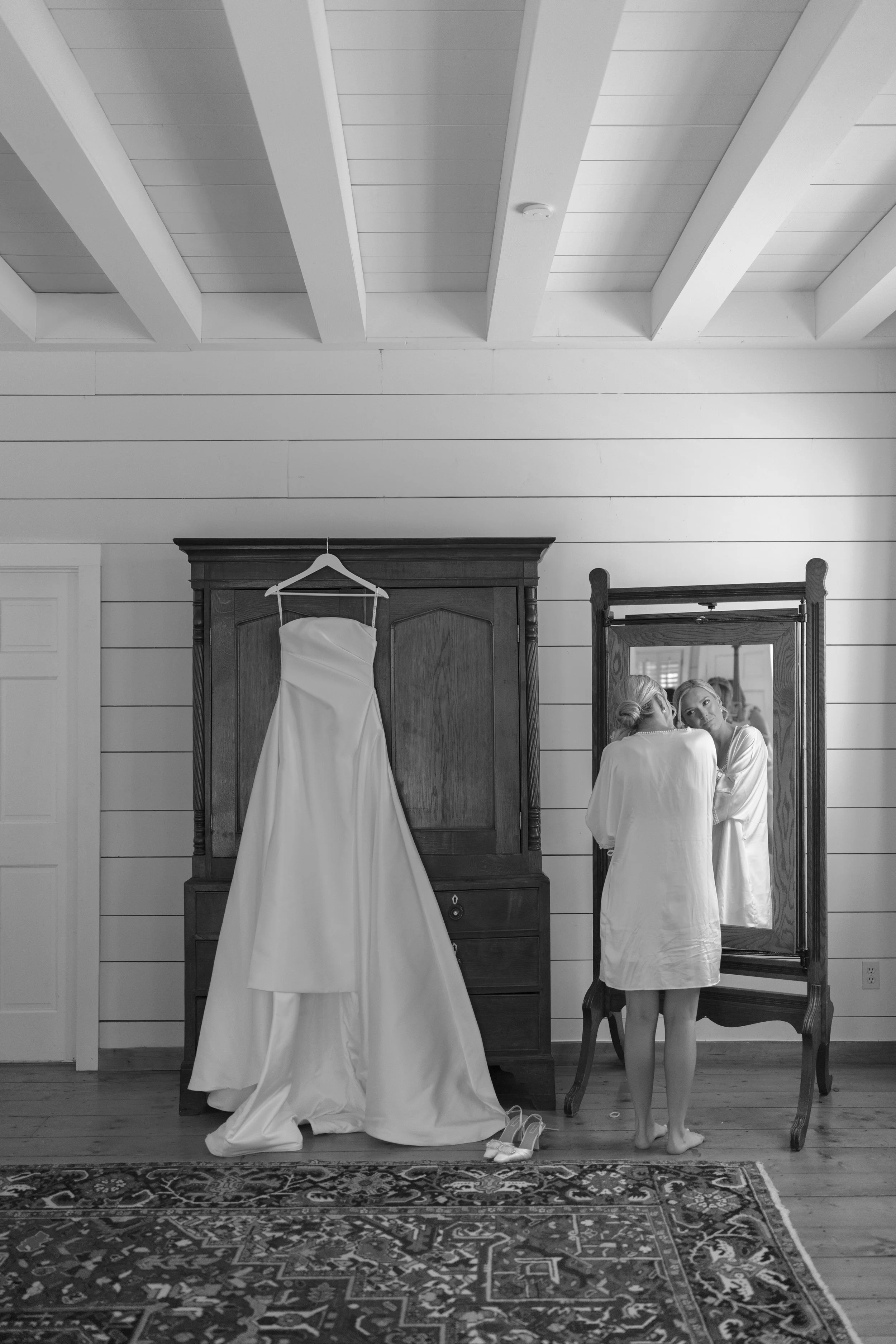 Black-and-white bridal dressing room scene with a wedding dress on a wardrobe, a bride reflected in a full-length mirror, and wedding shoes on the floor.