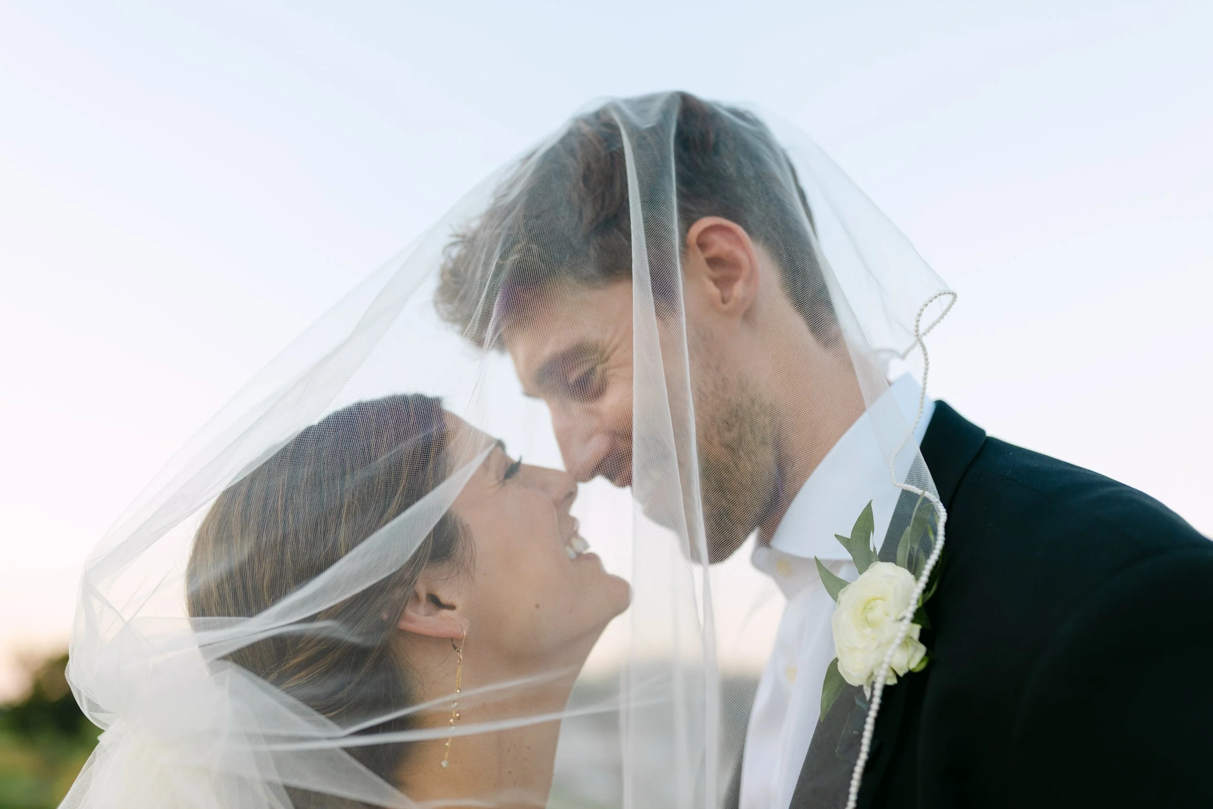 Bride and groom sharing a kiss under a veil outdoors during sunset.
