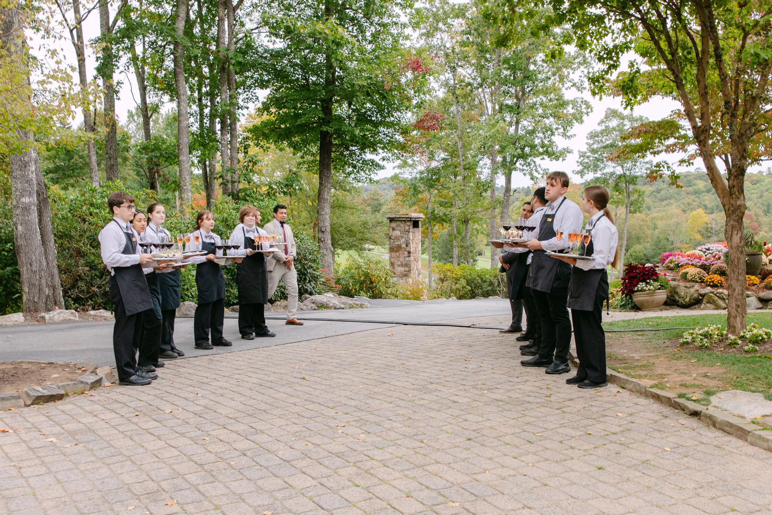 Catering staff in a landscaped garden holding trays of wine glasses during an outdoor event