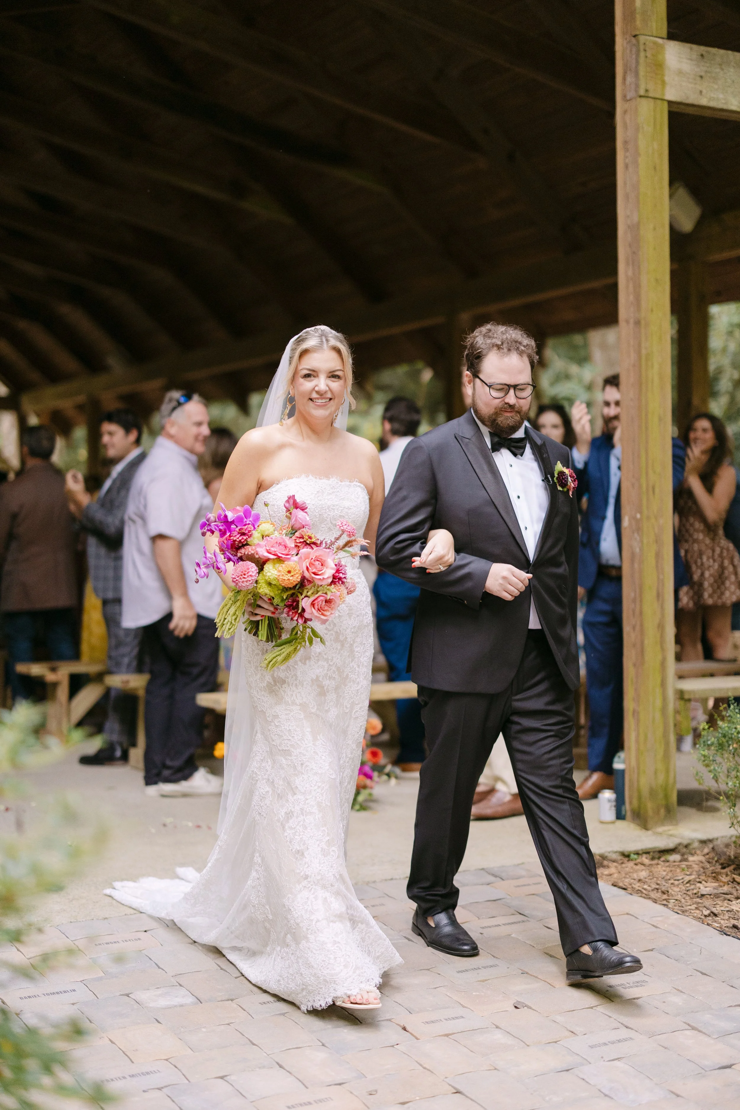 Bride in a white lace strapless wedding gown holding a colorful bouquet walking arm-in-arm with a groom in a black tuxedo during a wedding ceremony.