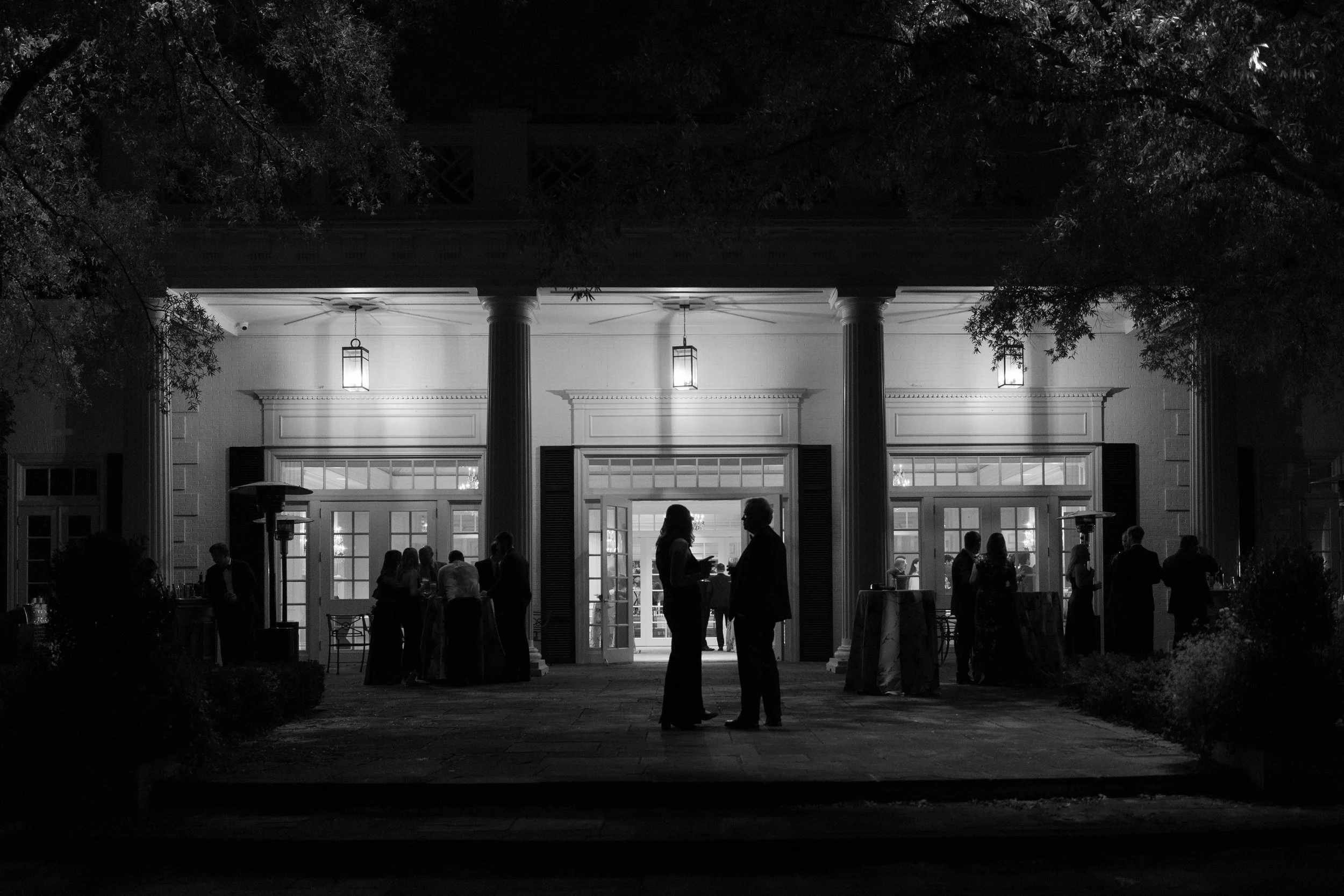 Silhouettes of people socializing outside a lit building at night, with trees and bushes framing the scene.