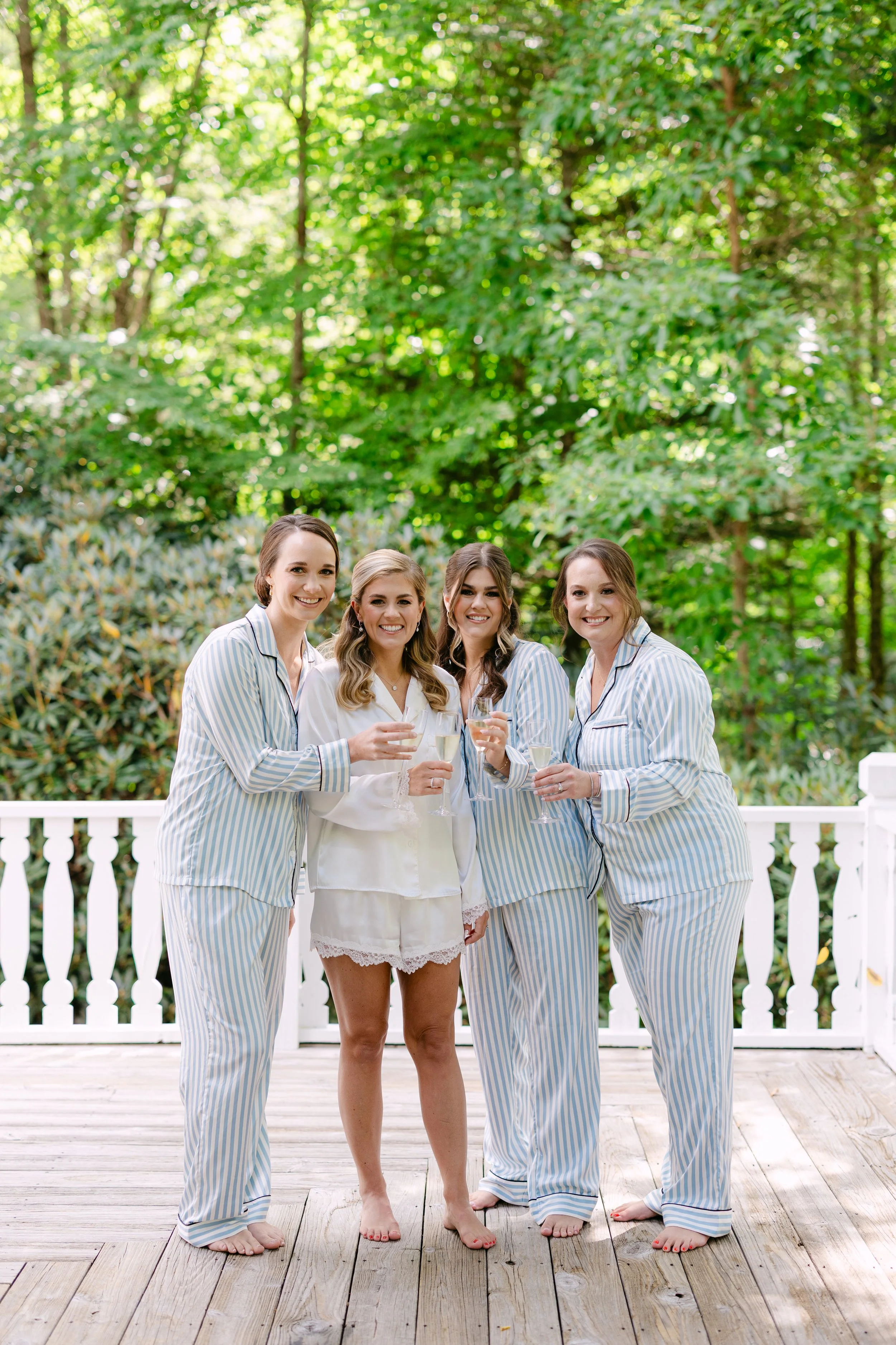 Group of four women in pajamas holding glasses of champagne on a wooden porch with lush green trees in the background.