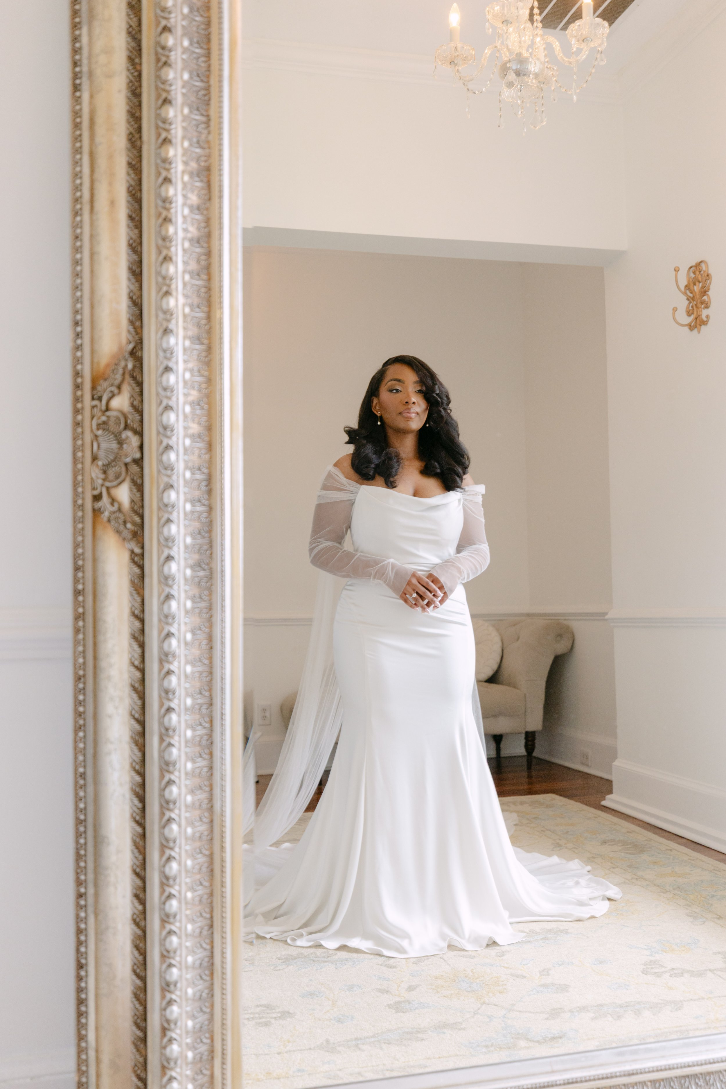 A woman in a white wedding dress standing and looking at her reflection in a full-length mirror in a room with cream-colored walls, a chandelier hanging from the ceiling, and a vintage-style cream-colored armchair in the background.