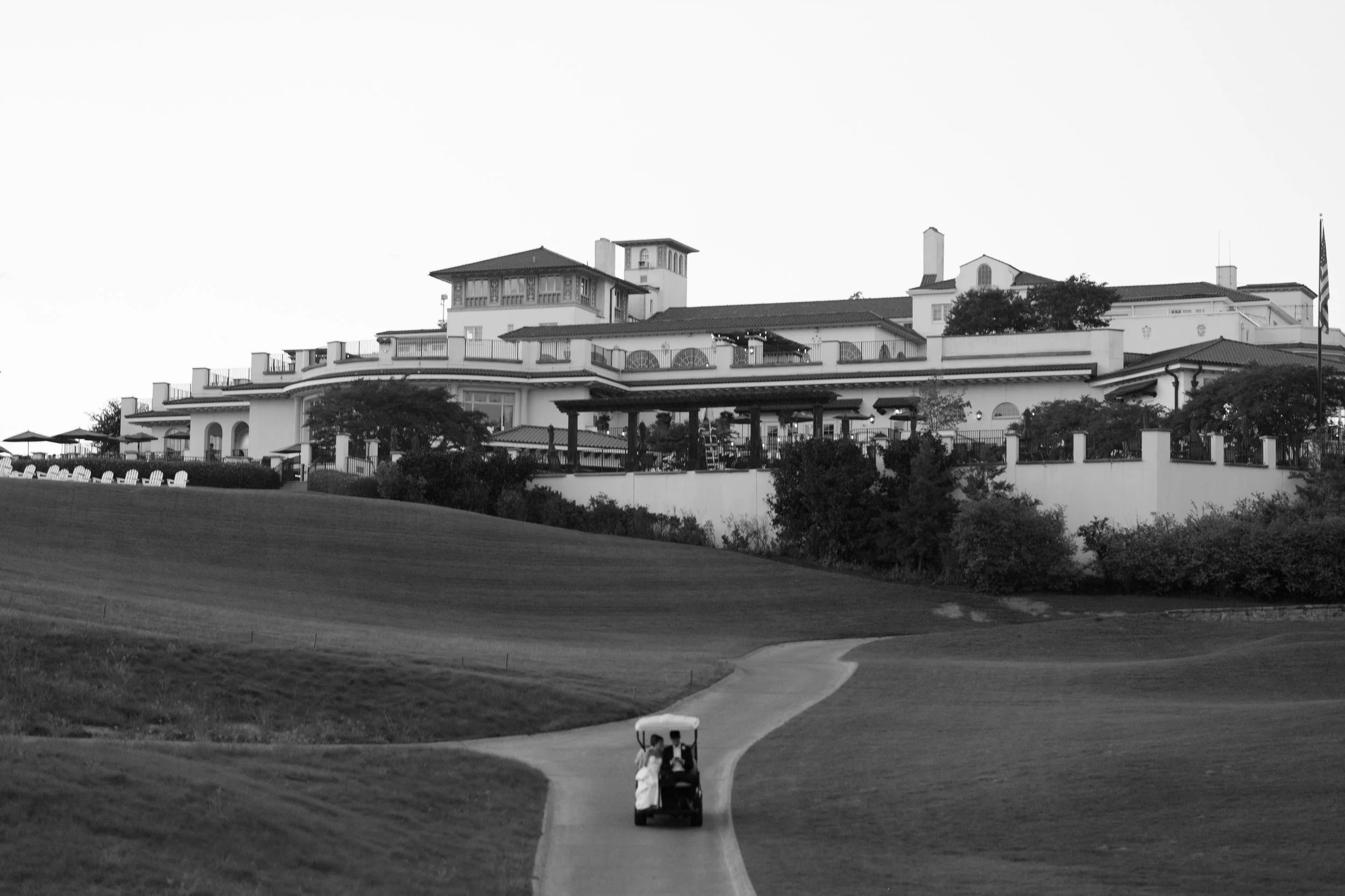 A golf course with a cart on the path, and a large building in the background, possibly a clubhouse, in black and white.